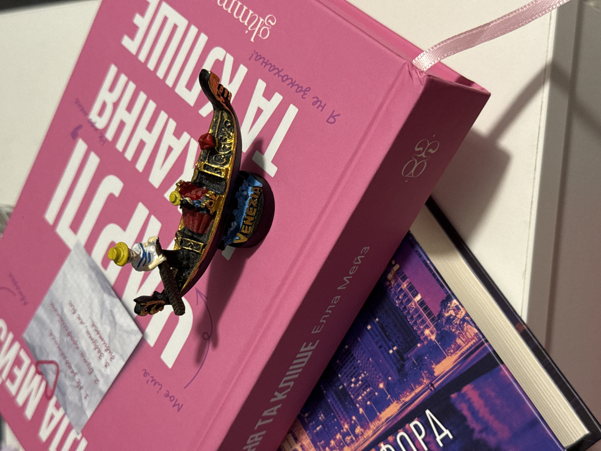 The image shows a close-up of two books, one pink and one with a dark cover featuring a cityscape. The pink book has large white text that appears to be a title. A small figurine of a gondola with two figures and a gondolier, along with a small piece of paper with handwritten text and a red heart-shaped paperclip, are placed on the pink book. The gondola figurine has "VENEZIA" written on its base. A pink ribbon bookmark hangs from the side of the pink book. The scene is lit from above, casting shadows on the surface. There are no identifiable landmarks or elements that can place the location in Łódź, Poland, or any specific city. The image is a still life composition of books and decorative items.