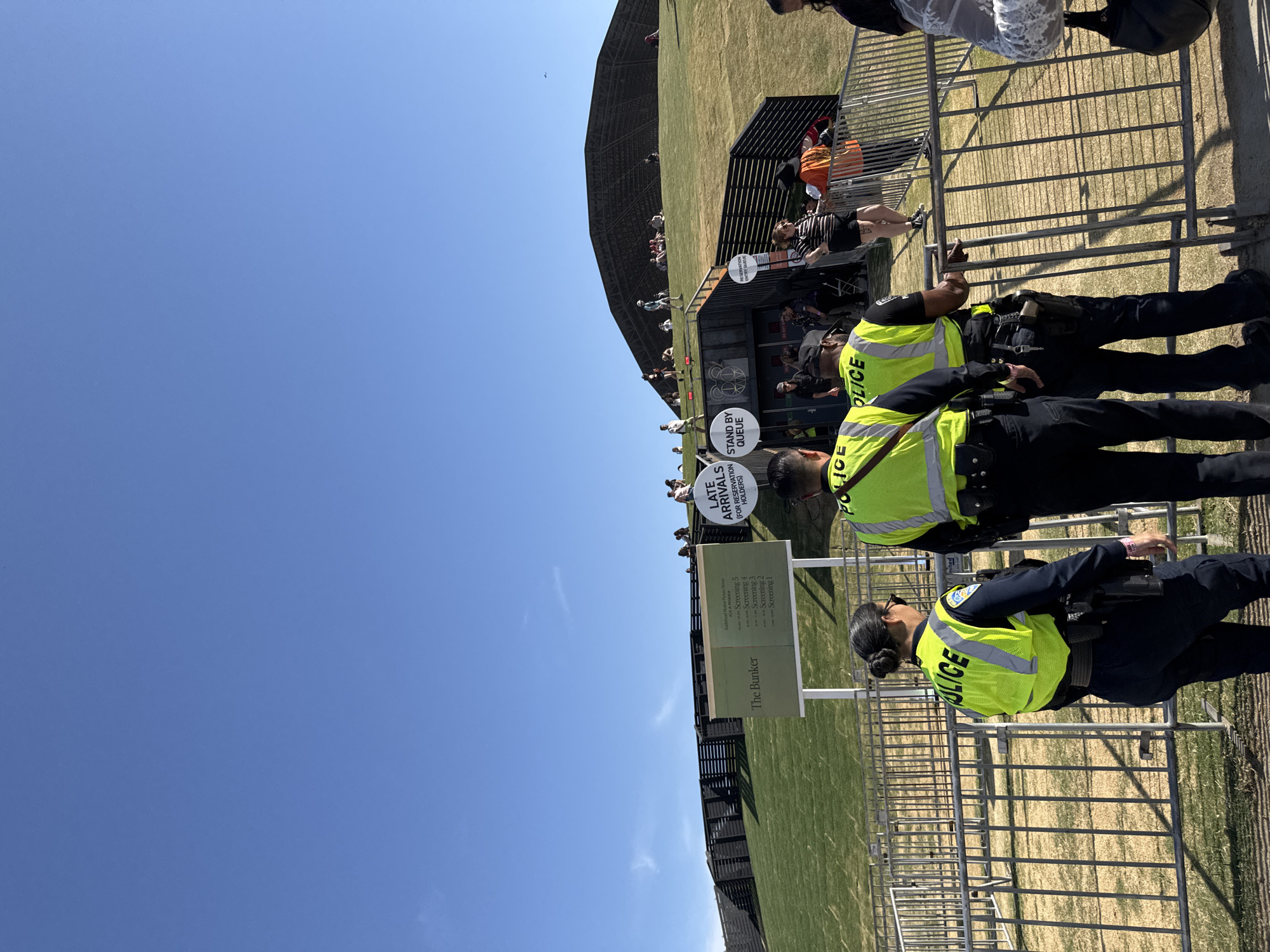 The image, taken outdoors in Indio, United States of America, shows a sunny day with a large grassy field adjacent to a dark-colored structure resembling an open-air pavilion or bunker, labeled "The Bunker" in a sign.  The sign also lists "Radiohead Motion Hota!" and "A Media" along with screenings numbered 1-5. A sign beside it says "LATE ARRIVALS (FOR RESERVATION HOLDERS) STAND BY QUEUE"

Three police officers, wearing neon yellow vests with the word "POLICE" printed on them and utility belts including what appears to be a firearm, are standing in the foreground near a metal barrier. People are queuing or sitting within the structure and along the grassy area. A dark fence runs parallel to the structure's perimeter. The sky is clear blue.