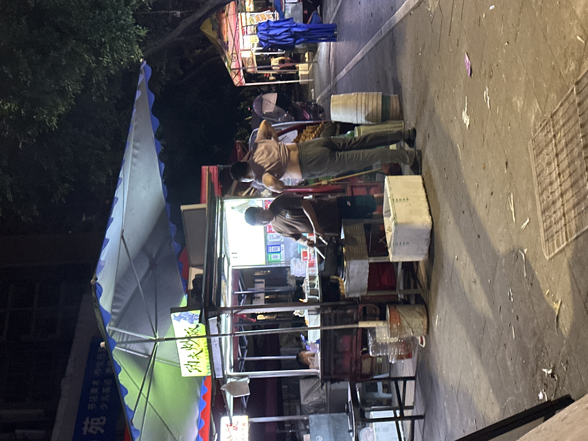 This nighttime scene, located in the Chongqing High-tech Zone, China, captures an active street food stall. A large, white and blue scalloped umbrella shades the primary food preparation area, illuminated from beneath by a green light. A yellow sign bearing the Chinese characters "功夫炒饭" (Gongfu Fried Rice) is prominently displayed.
At the main stall, an adult male in a dark brown t-shirt and dark shorts is actively cooking in a wok on a gas stove. To his right, another adult male, with his back partially turned, is adjusting a light-colored t-shirt, pulling it up to expose his midsection, while wearing dark pants. A third individual's head is partially visible behind the stall's counter in the background.
The immediate ground area is paved and contains several objects: a large white styrofoam cooler, multiple plastic bottles, and industrial buckets (one white, one orange). A light-colored, vertically stacked barrel stands near the man adjusting his shirt. Further down the street, to the right, another brightly lit stall with a multi-colored awning is visible. Next to this background stall, a tall, cylindrical object draped in blue fabric stands upright. Dark trees form the upper right background. The paved ground shows scattered small debris and discarded items. A partial view of a scooter with a person holding an umbrella is discernible in the midground between the central figures and background stalls. Artificial lighting casts distinct shadows across the scene.