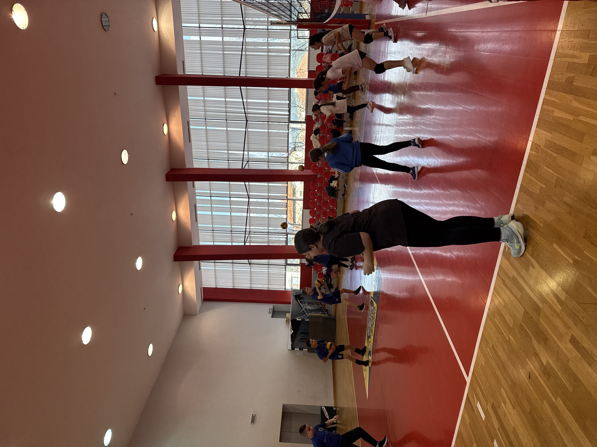 A gymnasium in Etropole, Bulgaria, features a varnished wood floor and a maroon-painted interior. Large windows with horizontal slats line one side of the room. A group of young people are doing warm-up exercises. A volleyball net is set up. Red chairs are positioned against the wall. Overhead, evenly spaced circular lights illuminate the scene.