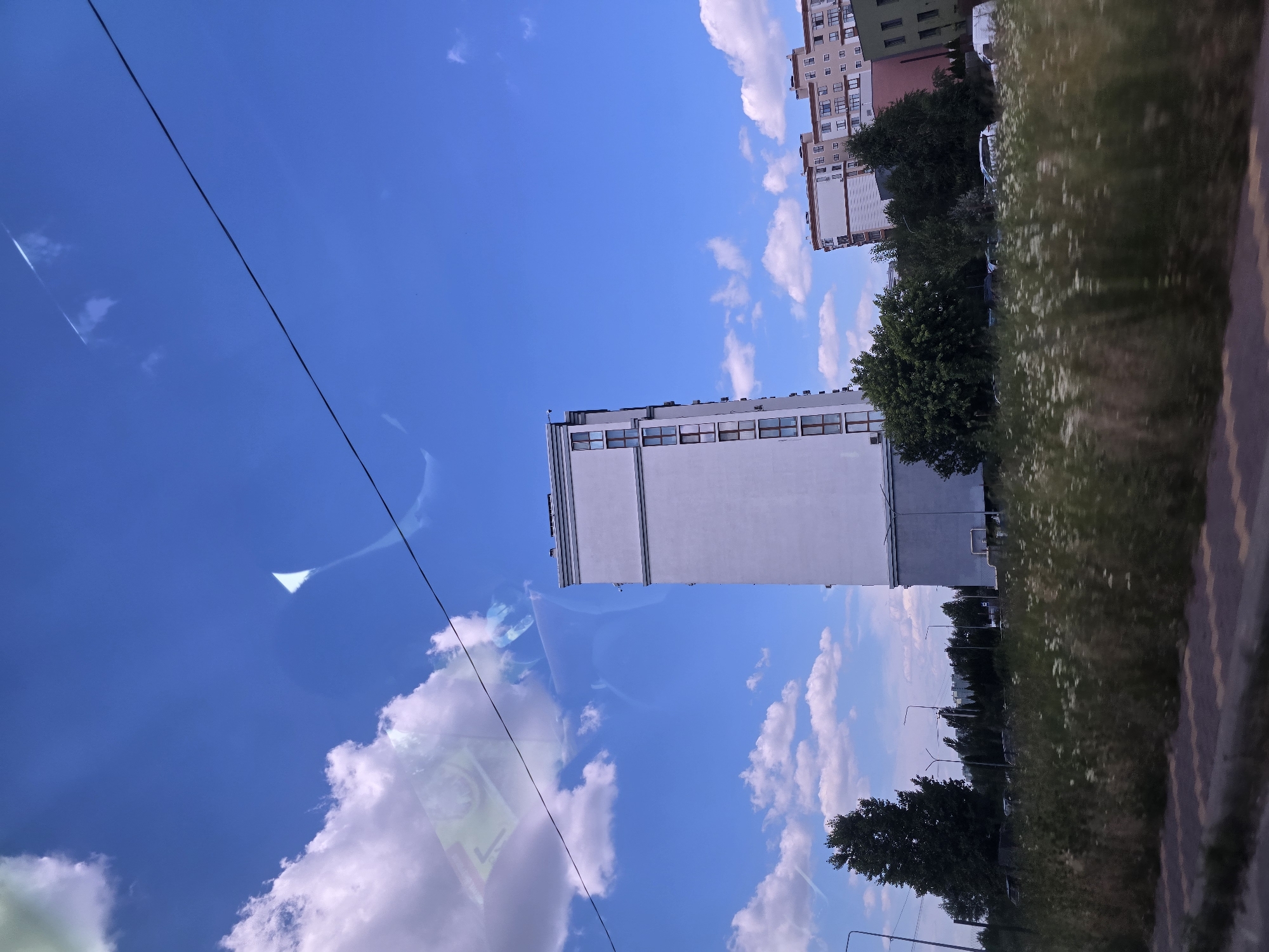 A tall, modern apartment building with a white facade and numerous windows stands prominently against a bright blue sky dotted with white clouds. To the right of this building, other apartment buildings of varying heights and colors are visible. In the foreground, a field of tall, green grass and wildflowers stretches across the bottom of the image, with a blurred asphalt road running along the very bottom edge. Trees are scattered throughout the scene, some larger and more mature, others smaller and more numerous, creating a lush, green backdrop. The overall impression is of a suburban or semi-urban environment in Petropavlivska Borshchahivka, Ukraine, bathed in daylight. There are no people visible in the image.