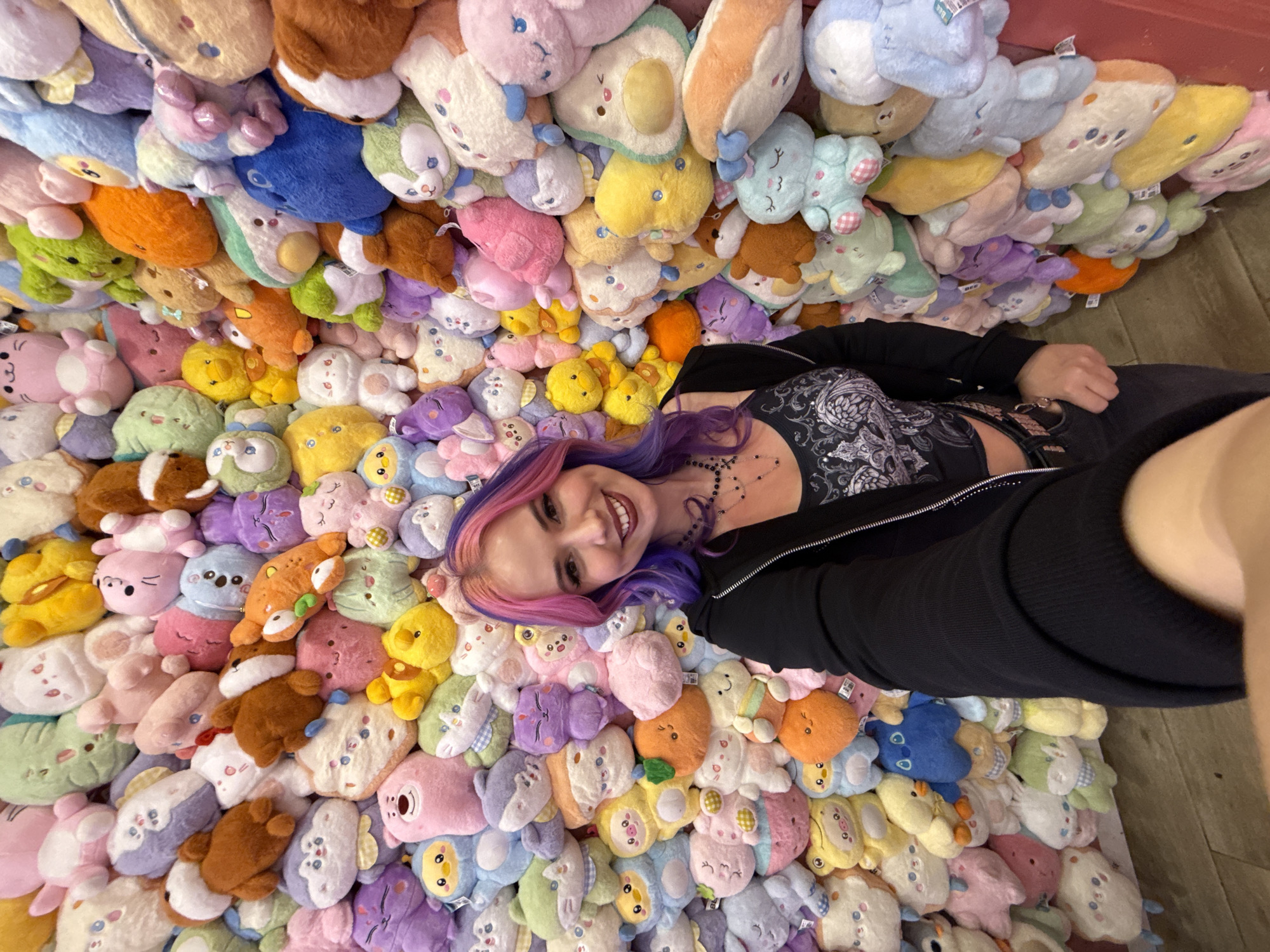 A selfie taken from above shows a smiling person lying among a dense layer of colorful plush toys. The person has hair dyed with pink and purple hues. They are wearing a black jacket and a dark top with a pattern. The plush toys vary in shape and color, including pastels and brighter shades, with some resembling animals and others more abstract shapes. The toys create a cushioned surface around the person, filling the frame entirely. There is no discernible evidence of Madrid, Spain.