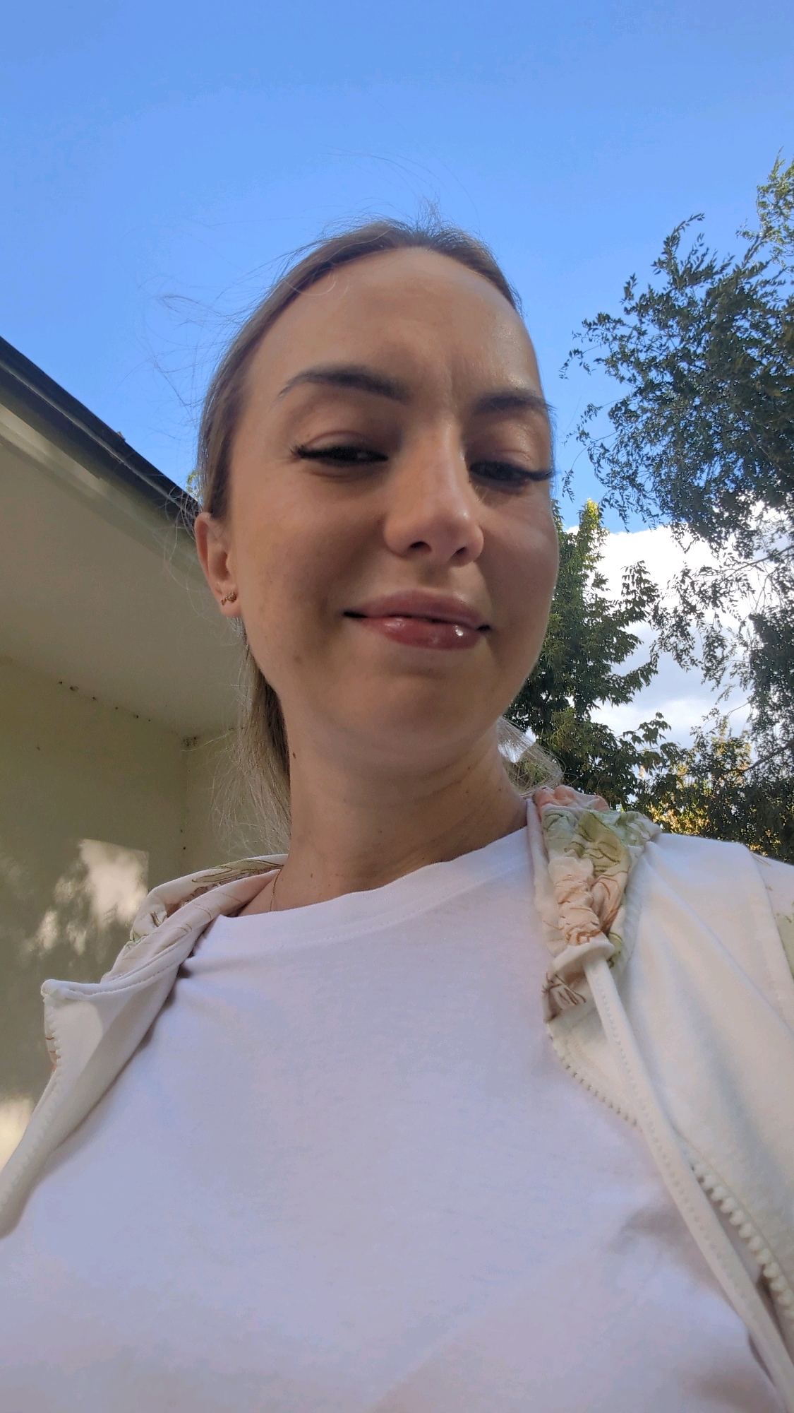 A young Caucasian woman is captured in a selfie taken outdoors. She is looking towards the camera with a slight smile. Her hair is pulled back, and she is wearing a white t-shirt and a light-colored jacket with a floral pattern on the collar and cuffs. The background features a clear blue sky with some wisps of clouds, and the top of a building with white siding is visible on the left. Trees with green foliage are present on the right. The overall scene suggests a casual outdoor setting, potentially a residential area or park. There is no information in the image to suggest a specific location within Chișinău, Moldova.