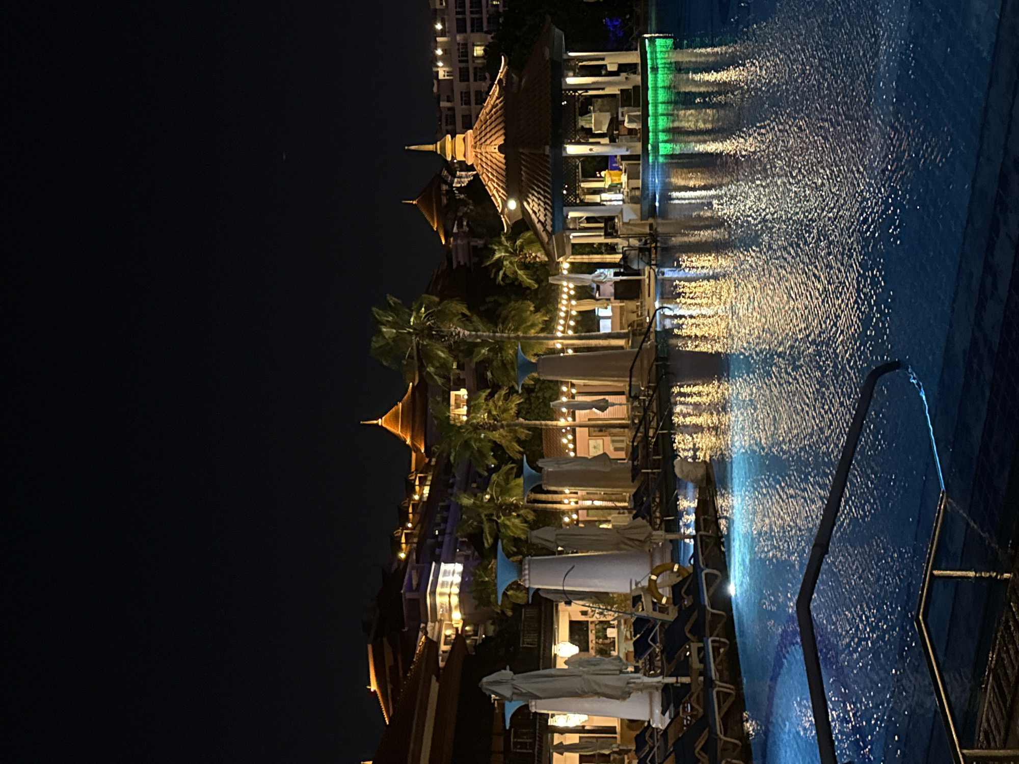 The image depicts a nighttime scene at a resort in Dubai, United Arab Emirates. A large swimming pool occupies the foreground, with its surface illuminated by underwater lights and reflections of surrounding structures. In the mid-ground, a swim-up bar with a thatched roof is prominently featured, glowing with green lights. Lounge chairs and umbrellas are visible along the edge of the pool and surrounding deck, indicating a recreational area. Palm trees and resort buildings, characterized by their distinctive architecture and warm lighting, form the background. The overall atmosphere is one of a luxurious and tranquil evening setting.