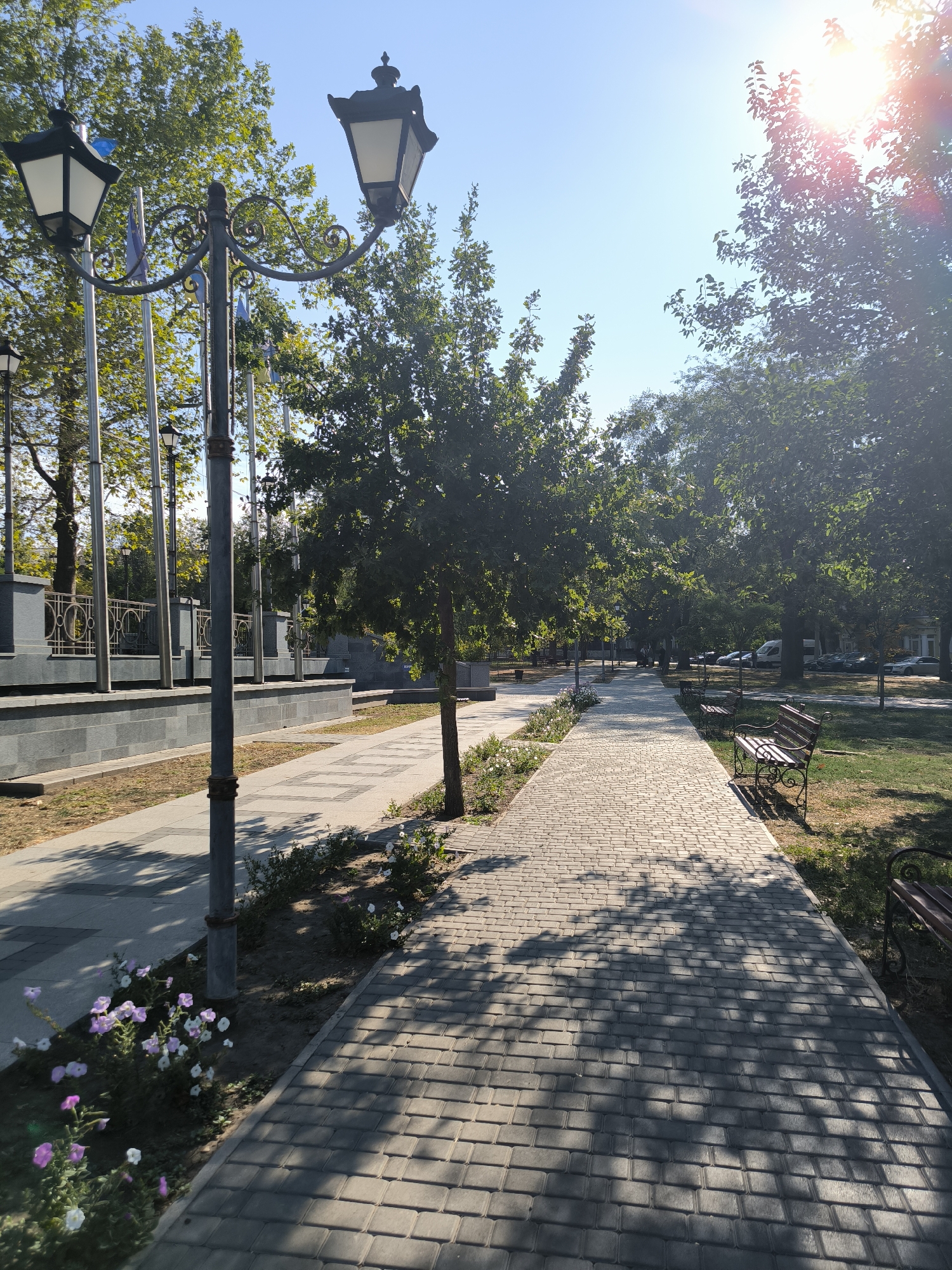 The image depicts a paved pathway in a park-like setting in Mykolaiv, Ukraine. The pathway is constructed with light-colored interlocking pavers, casting dappled shadows from the overhead trees. To the left of the pathway, there is a raised grey stone structure with a decorative metal railing, and several tall flagpoles. A decorative streetlamp with two lights stands prominently in the foreground on the left. Small flowering plants with white and purple blossoms are visible along the edge of the pathway. On the right side of the pathway, there are two wooden park benches with ornate metal legs, positioned on a grassy area. Mature trees with full green foliage line the pathway and the park area, and the sun is shining brightly, creating lens flare in the upper right corner of the image. In the background, a road with several parked vehicles can be seen. The overall scene suggests a well-maintained public space on a sunny day.
