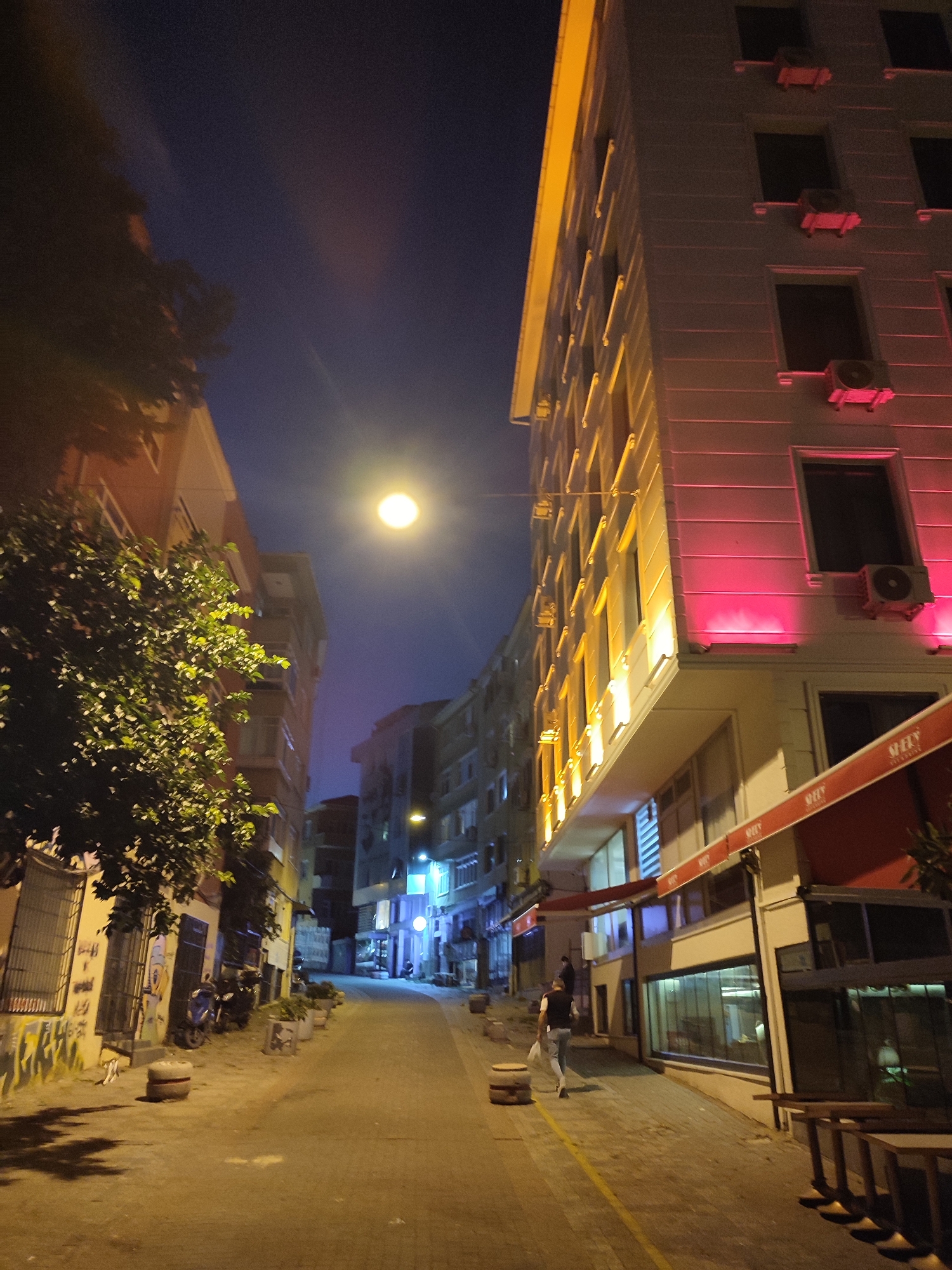 The image shows a street scene in Kadıköy, Turkey, at night. The sky is dark blue with a bright, luminous full moon. On the right side of the street is a modern, multi-story building with its facade illuminated by pink and yellow lights. Several air conditioning units are visible on the exterior walls. Red awnings are extended over the ground-floor windows. A person is walking down the street away from the camera.
On the left side of the street, there is an older building with windows and some trees in front. The street is paved with cobblestones and there are several concrete planters lining the sidewalk. A few parked motorcycles are visible against the wall of the building. The overall atmosphere is urban and nocturnal, with artificial lights creating a dramatic effect.