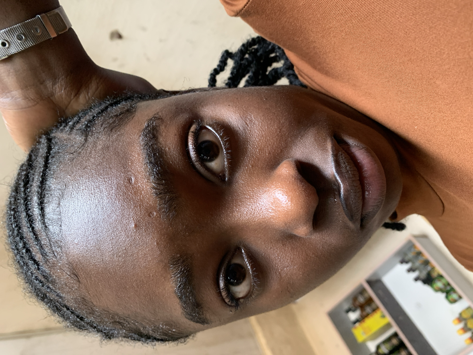 This image depicts a close-up portrait of an individual, likely situated indoors in New Stadium, Nigeria. The subject, a person with dark skin, looks directly into the camera. Their hair is styled in cornrows, and visible baby hairs frame the forehead. The skin exhibits a luminous sheen, particularly on the forehead, nose, and lips, indicating direct lighting. Small skin texture variations and minor blemishes are visible on the forehead. Both dark brown eyes are open, with visible eyelashes and subtle light makeup lining the lower waterlines. The lips are full and appear lightly outlined. The subject's right hand and wrist are visible in the upper right, positioned near the head, adorned with a silver-toned watch featuring a metallic mesh band with circular cutouts and a rectangular clasp. An orange-brown, ribbed garment is worn. In the blurred background to the lower left, two multi-compartment organizers contain numerous small bottles of varying colors (amber, green, clear) and caps, alongside a yellow box. The remainder of the background consists of a plain, light-colored surface.
