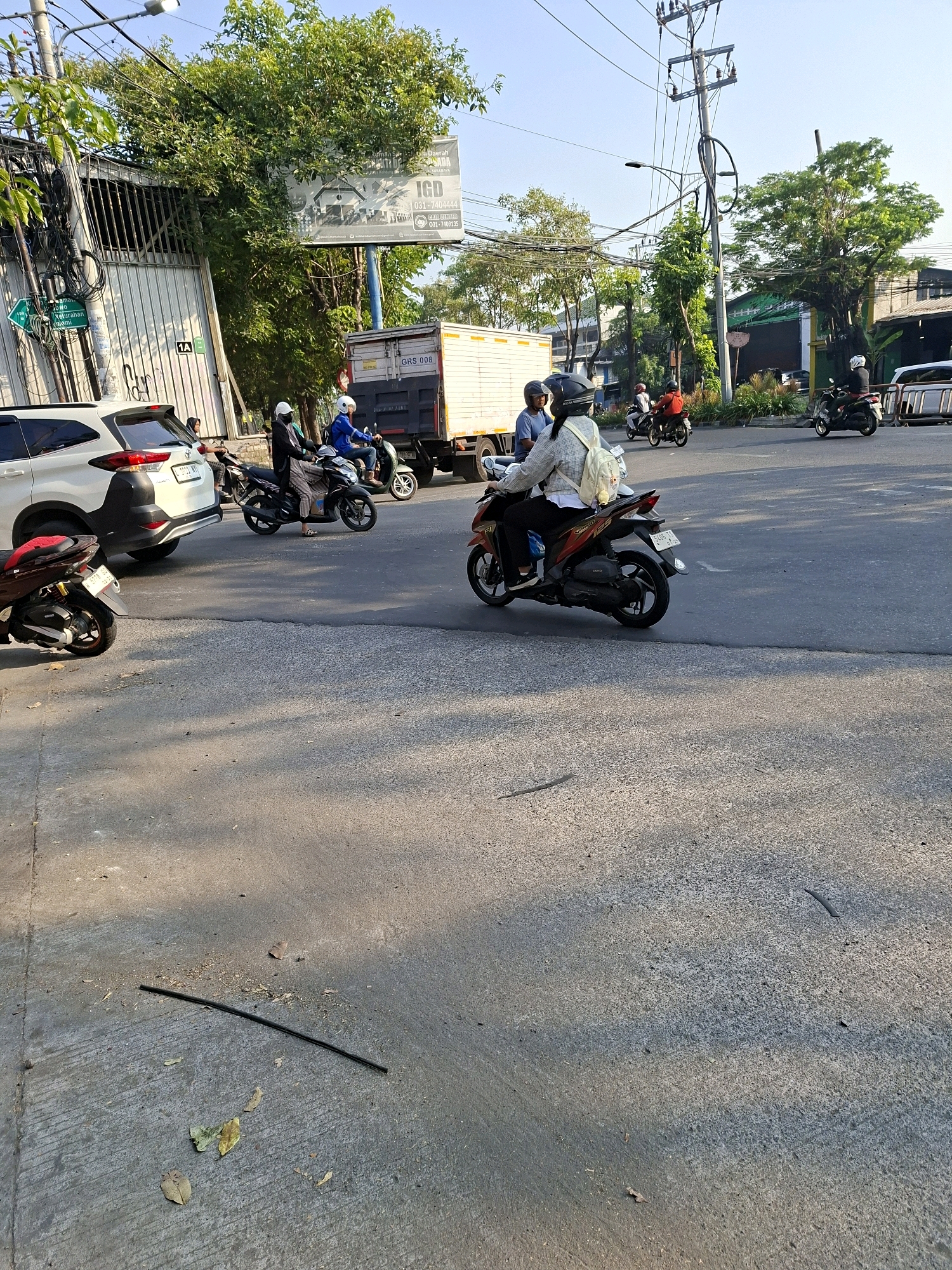 This image captures a street scene in Surabaya, Indonesia, under a clear sky during daylight. A white SUV is parked on the left side of the road, with its rear towards the camera. A dark-colored motorcycle with a rider wearing a white helmet and a patterned shirt is in the foreground, partially obscuring the SUV. Several other motorcycles, some with riders, are visible further down the road, moving in the same direction. One motorcycle has a rider in a blue jacket and white helmet, and another has a rider in a red shirt and white helmet. A white truck with the marking "GRS 008" is stopped in the middle of the intersection. The road surface is asphalt, with some debris and fallen leaves scattered on the ground. A pole with numerous electrical wires and a streetlamp stands tall on the right side of the frame. In the background, there are buildings and lush green trees, suggesting an urban environment. A billboard with text is visible on the left side of the image, mounted on a blue pole, with trees growing around it. The overall impression is a typical, busy street intersection in a city.