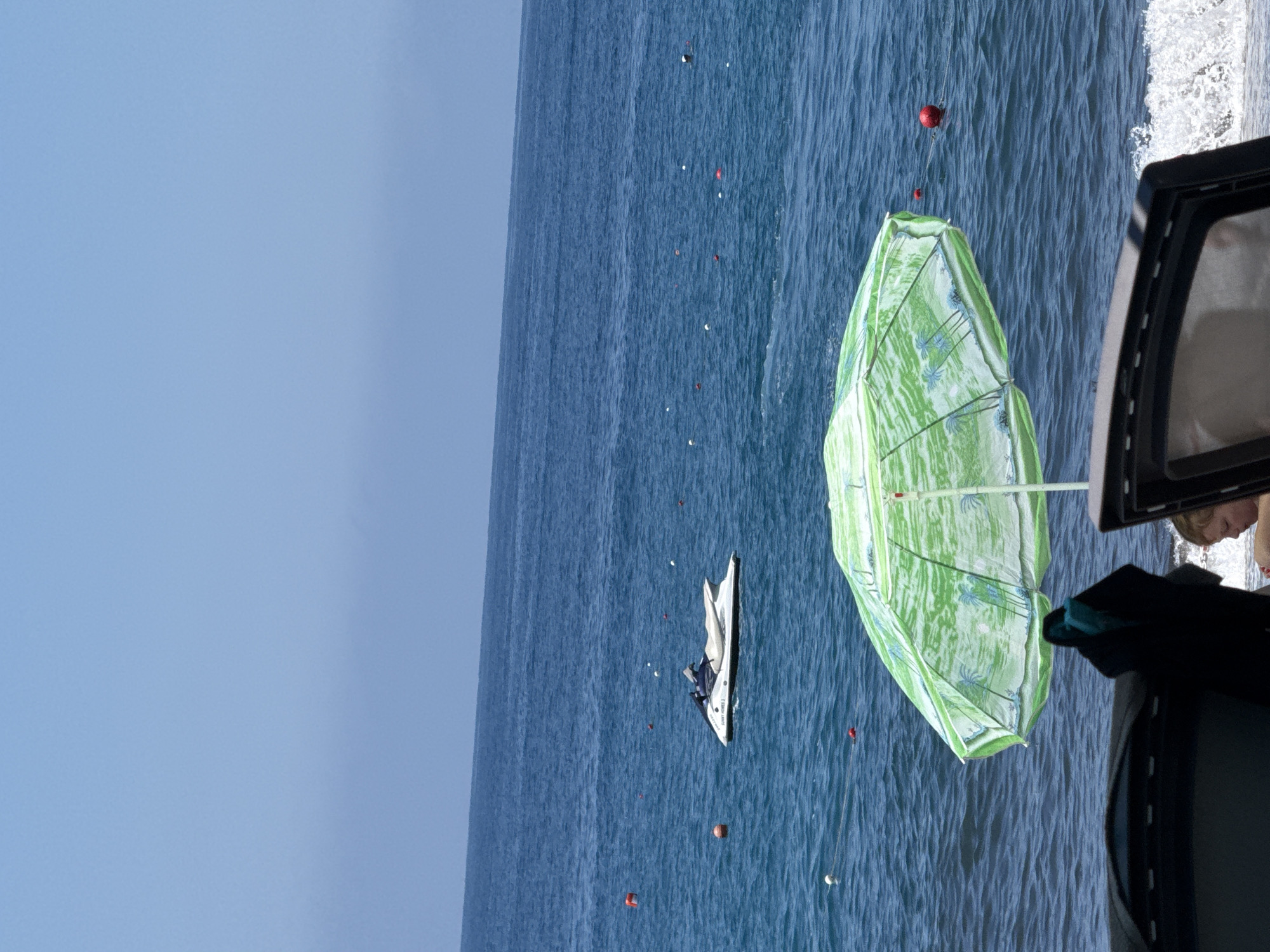 A wide-angle, upside-down shot captures a coastal scene, possibly near Unknown. The clear blue sky transitions to a vast expanse of dark blue sea, marked by numerous small, red floating objects. A white jet ski with a person on board is visible in the mid-ground. To the right, a green and white patterned umbrella partially obscures the view. A portion of a vehicle is also present, providing a partial view of a person with reddish lips inside. The photo appears to be taken from inside another vehicle.