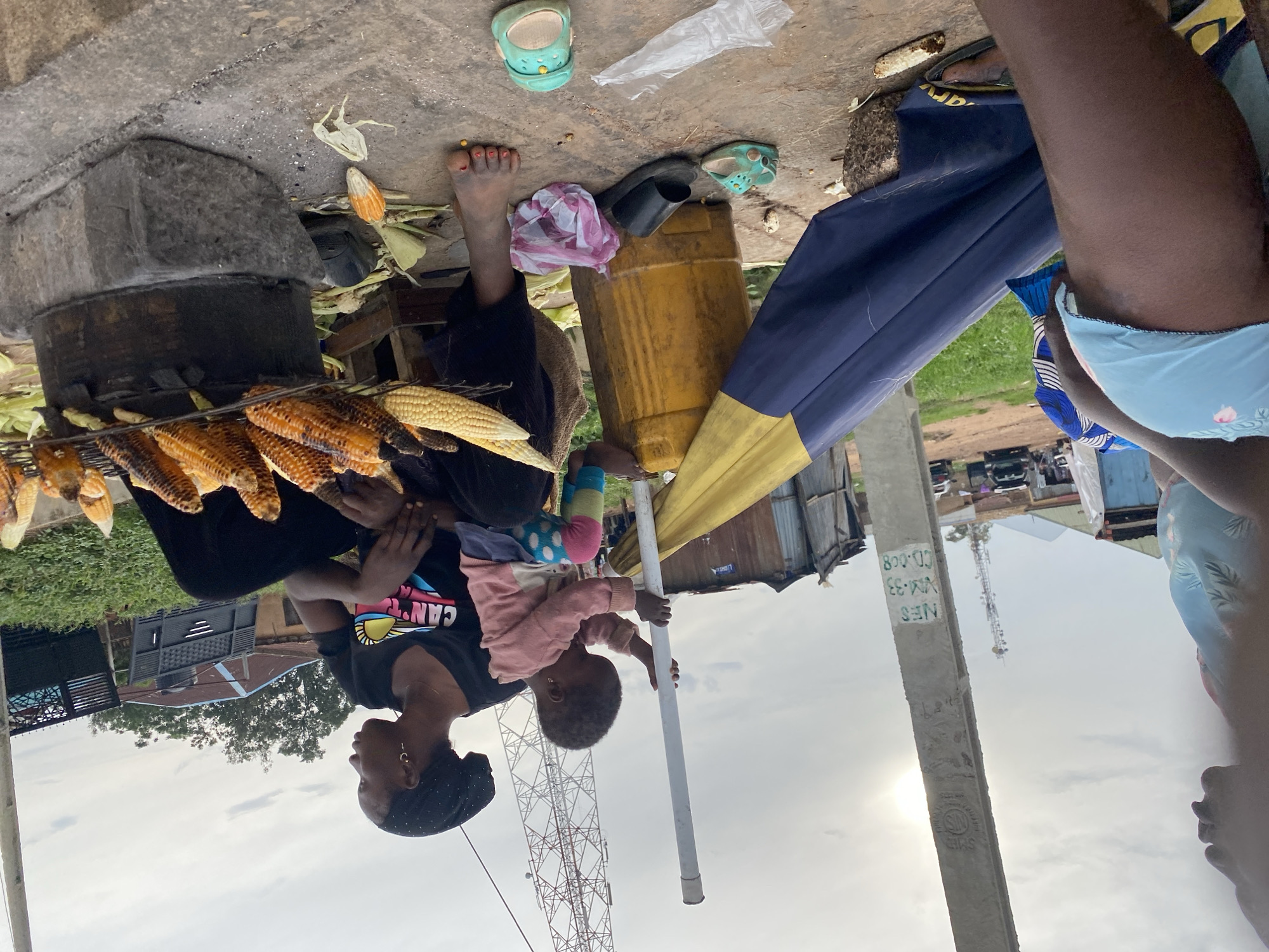 A street scene in Bukuru, Nigeria, shows a woman and child next to a charcoal grill used for roasting corn. The woman, wearing a black t-shirt and head covering, holds a young child wearing pink. The grill contains several ears of corn cooking. Beside the grill are various objects including a yellow container, a small black sandal, and a pink and white cloth. A utility pole is also visible, with text in white, which reads "NES 800.03 VM-33 CD-008 SMS". In the background is a cell tower, a building, and a vehicle. The scene appears to be taken outdoors, with a gray sky overhead.