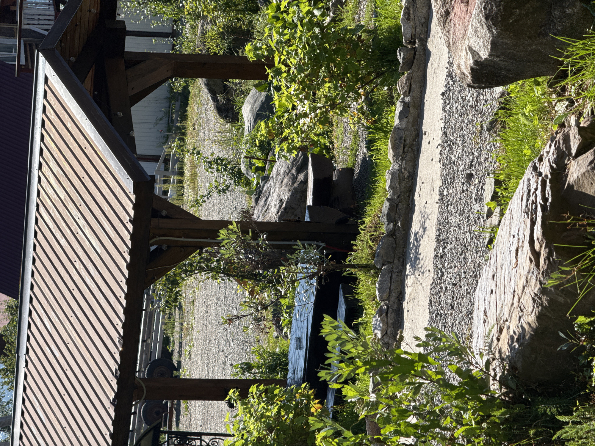 The image depicts an outdoor seating area with a wooden structure covered by a corrugated metal roof. Beneath the shelter, a dark-colored picnic table and benches are visible. The ground surrounding the area is a mixture of gravel and grass, with several large rocks scattered throughout. Lush green bushes and plants frame the scene, and a stone border runs along a pathway. In the background, a trailer with wheels is partially visible. The scene appears to be situated in a residential or recreational setting. No individuals are present in the image.