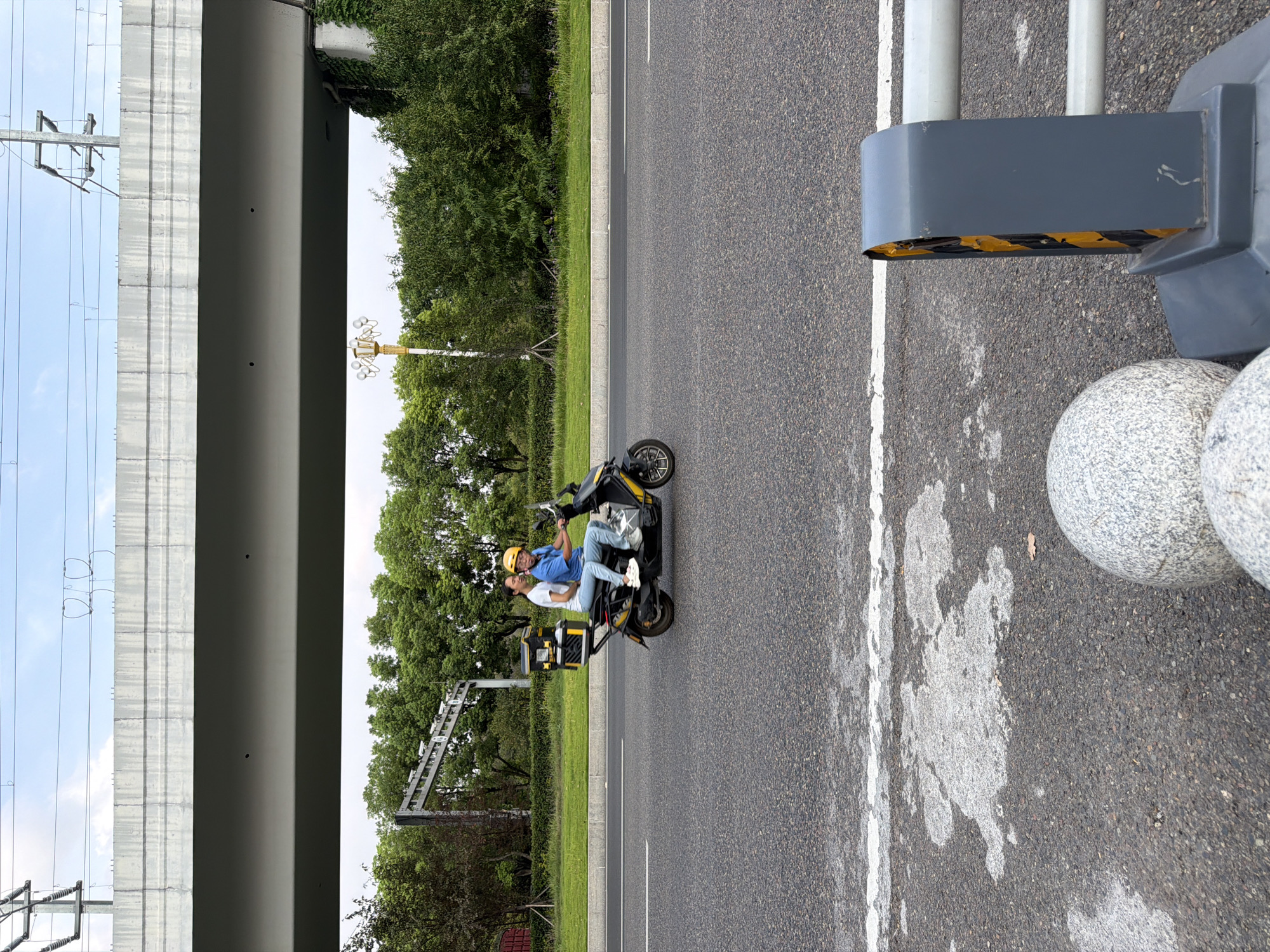 In Jiangjin, China, two individuals, a male driver and a female passenger, travel on a scooter under an elevated highway. The driver, wearing a blue shirt and yellow helmet, operates the scooter. The passenger, in a white shirt and wearing a white helmet, sits behind him. The scooter is equipped with a black cargo box mounted on the rear. The road surface is asphalt with white lane markings. A concrete structure supporting the highway spans the upper portion of the image, with power lines visible above it. Lush green trees and vegetation line the side of the road, and a street lamp with multiple bulbs stands in the background. In the foreground, a gray traffic barrier with yellow and black hazard stripes is visible, along with two large, round, granite-like decorative spheres. The scene depicts an ordinary moment of transportation in an urban environment.