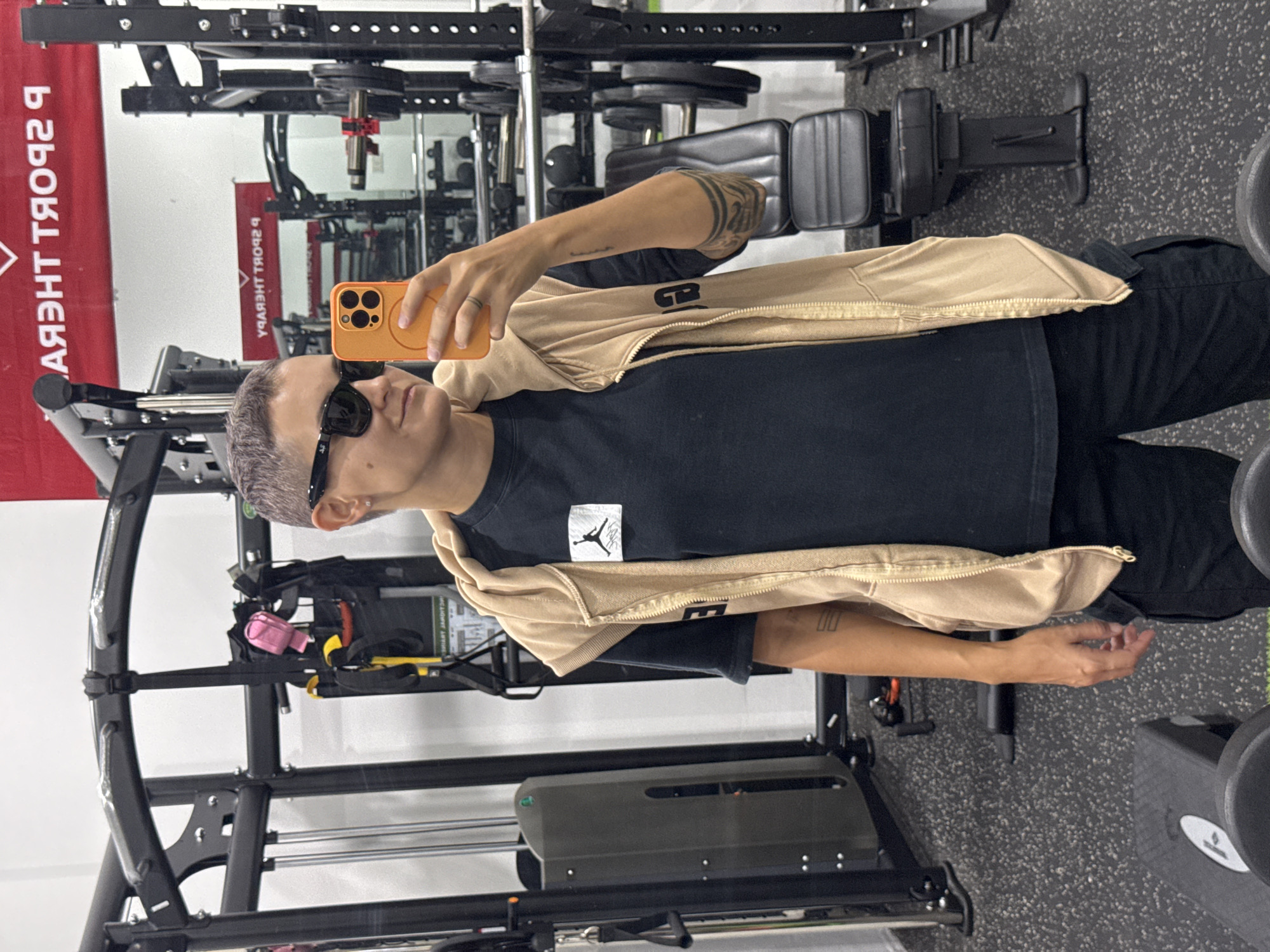A person with short, gray hair and sunglasses is taking a selfie in a gym. They are wearing a black t-shirt with a white logo and a tan hooded vest. The person is holding an orange smartphone in their right hand. In the background, there is gym equipment, including a weight rack, a bench, and a cable machine. The gym appears to be well-lit and clean. The person is standing on a black rubberized floor. There is a red banner visible in the background with text that appears to be in a foreign language. The location of the gym is not specified in the image, but the request asks to assume it is in Nam Nha Trang Ward, Vietnam.
