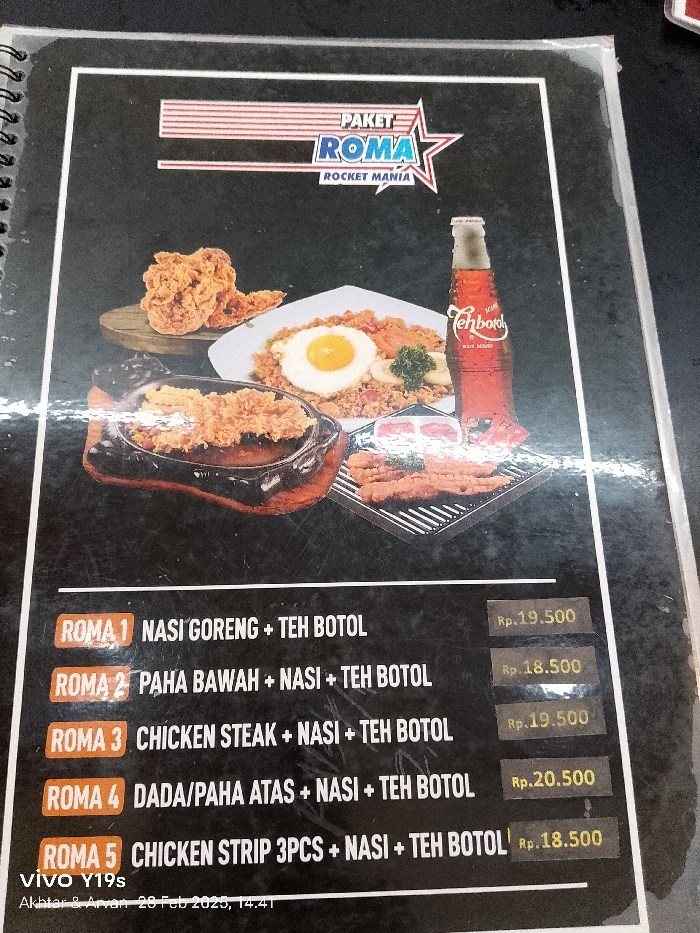 The image shows a menu board from a restaurant called "Paket Roma Rocket Mania" located in Patrol, Indonesia (latitude: -6.318, longitude: 107.986). The menu lists five different food options, each containing fried chicken, rice and a bottle of Teh Botol sweet tea, with varying prices ranging from 18,500 to 20,500 Indonesian Rupiah. The menu board is made of black and white cardboard with colorful text and stickers for the prices. The board is displayed in a vertical position. Above the menu board, there are images of the food options with the fried chicken, rice, and a bottle of Teh Botol. The images are colorful and glossy.