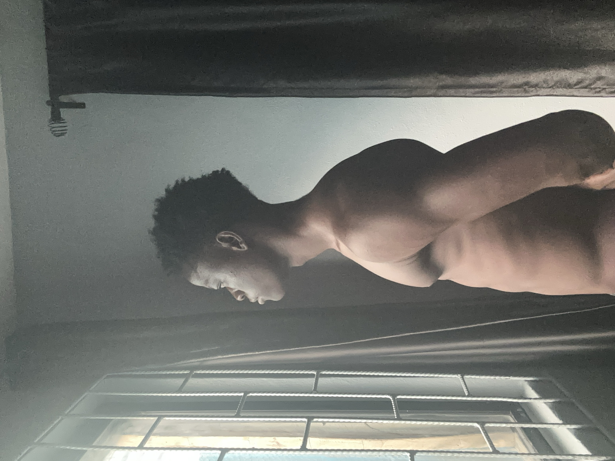 A dark-skinned young man with short, dark, curly hair stands in profile facing left, looking out a window with metal bars. He is shirtless, revealing a lean torso. The background consists of a wall and dark curtains. The window is partially obscured by the man and the curtains. The scene appears to be indoors, possibly in a room within a building in Badagry, Nigeria.