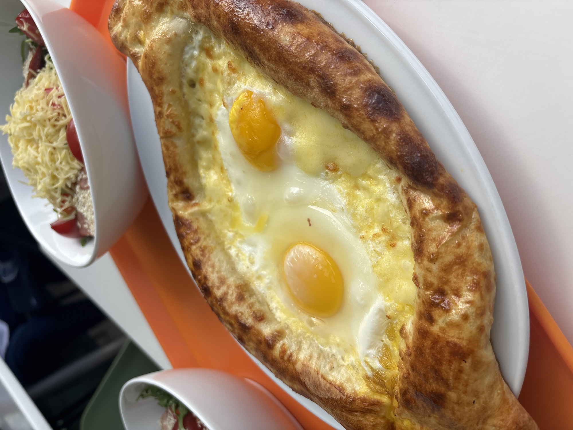 An eye-level, slightly angled, close-up shot displays a boat-shaped khachapuri, a traditional Georgian bread filled with cheese and two sunny-side-up eggs. The bread has a golden-brown crust. It is served on a white, oval-shaped plate. Adjacent to the khachapuri are two white bowls containing salad. The bowls are placed on an orange tray. The scene is lit by natural light. The setting is likely a restaurant in Yerevan, Armenia, based on the presence of traditional Caucasian cuisine.