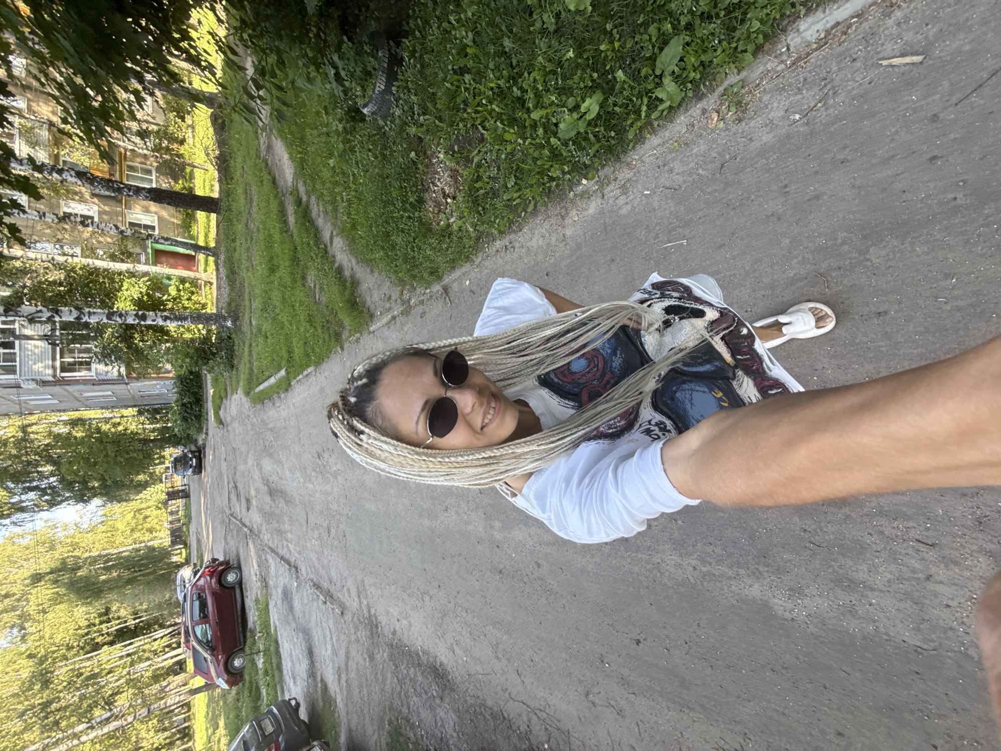 A woman with blonde braided hair and sunglasses is taking a selfie in what appears to be a residential area in Rusanovo, Russia. She is wearing a white t-shirt with a graphic print and white sandals. The background shows a dirt road, a parked red car, a multi-story apartment building, and trees. The lighting suggests it is daytime, possibly late afternoon due to the long shadows. The scene is consistent with a typical courtyard in a Russian town or city.