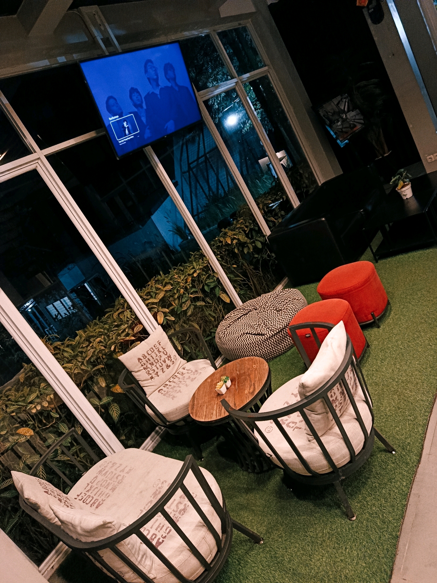 The image shows a modern seating area at night, likely in an indoor or semi-outdoor cafe or lounge in Tamansari, Indonesia. A large flat-screen television is mounted on a wall, displaying a group of people and text in a dark blue hue. The seating area is furnished with a patterned beanbag chair, two round red ottomans, and two armchairs with metal frames and light-colored cushions. The cushions have a stencil-like print of letters and numbers. A small round wooden coffee table with a decorative top sits between the armchairs. The floor is covered with artificial green turf. Behind the seating area, through large windows with white frames, lush green foliage is visible, suggesting a garden or outdoor space. The overall ambiance is contemporary and relaxed, with the lighting indicating nighttime.