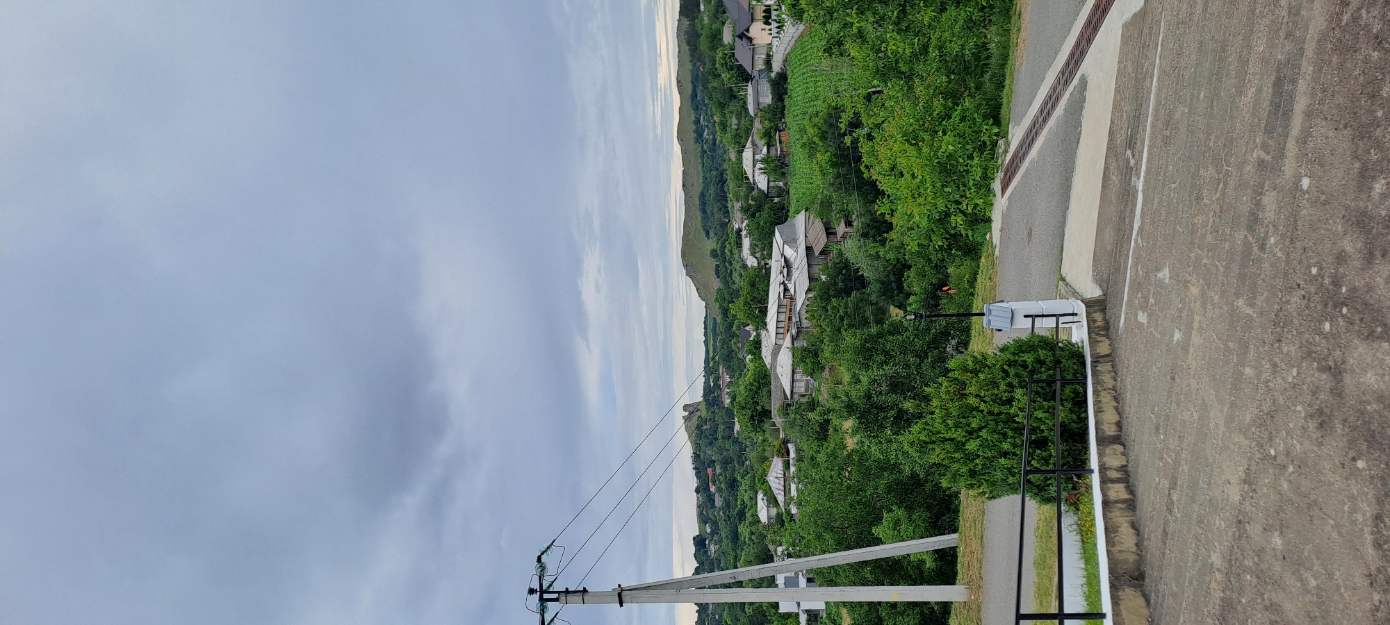 The image presents a wide shot of the village of Corjeuți, Moldova, under a cloudy sky. In the foreground, a concrete staircase leads down from the viewer's perspective, with a small portion of a road visible to the left and a drain grate crossing a paved area to the right. A black metal railing runs along the edge of a raised flower bed planted with a small evergreen tree.
Dominating the mid-ground is a dense expanse of green trees and vegetation interspersed with numerous houses. The houses are of varying sizes and architectural styles, with many featuring pitched roofs. Some appear to be older, with weathered roofs, while others seem more modern. A prominent wooden electricity pole with insulators and wires stands to the left of the frame, adding an element of infrastructure to the rural scene.
In the background, rolling hills covered in greenery frame the village. On one of the distant hills, a distinctive rock formation or ancient structure is visible, adding a point of interest to the landscape. The overall impression is one of a quiet, somewhat overcast day in a rural Moldovan village. No people are visible in the image.