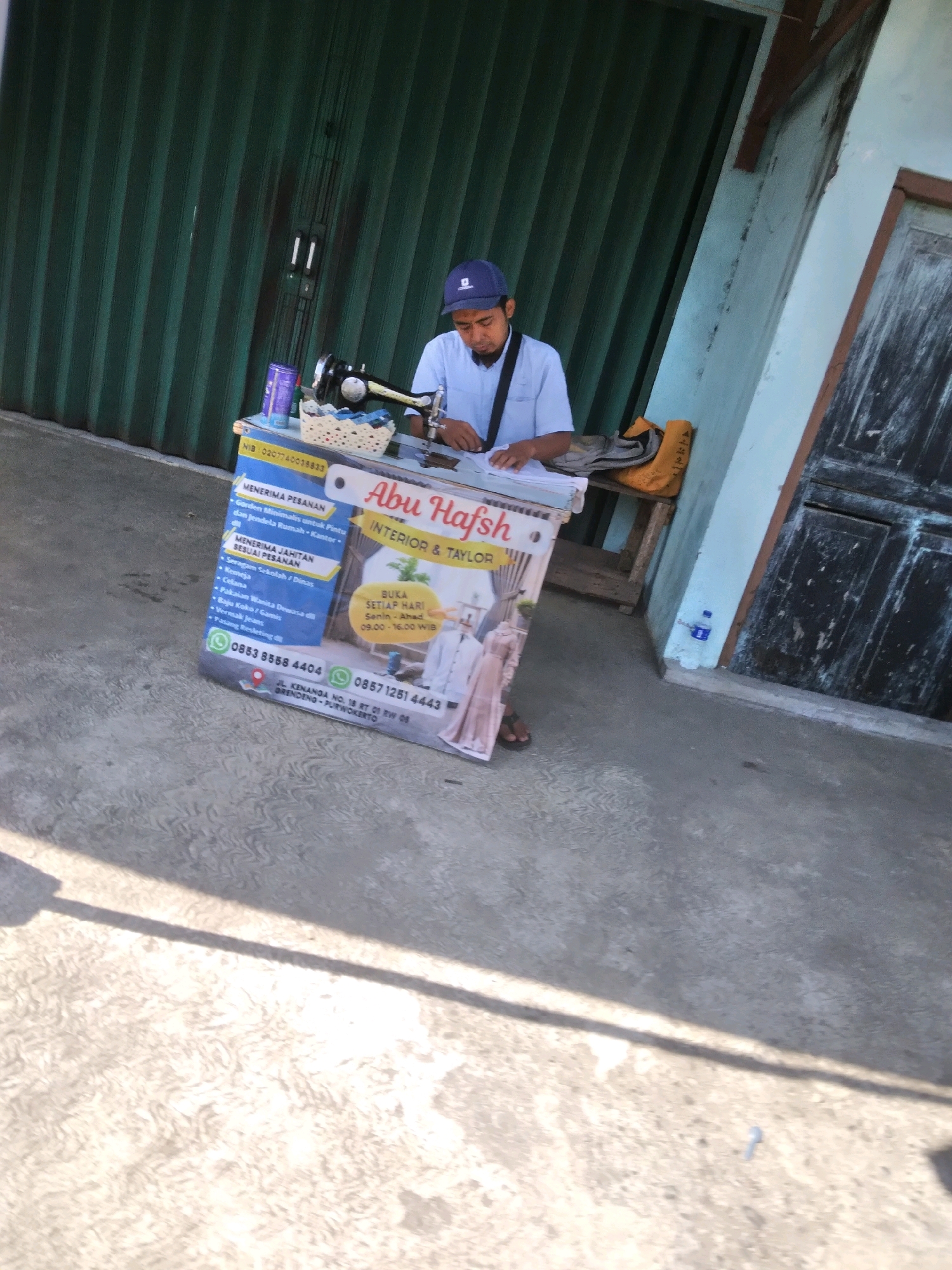 A man wearing a blue baseball cap and a light blue short-sleeved shirt stands behind a white and blue portable sewing counter. The counter displays advertisements for "Abu Hafsh Interior & Taylor" services. The man is operating a black sewing machine, with his hands on the fabric and machine. Next to the sewing machine is a small white woven basket. The background features a dark green corrugated metal door and a light blue wall. A yellow bag and a brown wooden stool are visible behind the man. The scene appears to be outdoors, with a grey concrete ground and a shadow cast across it. The advertisement on the counter lists services like "Menerima Pesanan" (Accepting Orders) for "Gorden Minimalis untuk Pintu dan Jendela Rumah Kantor" (Minimalist Curtains for Home and Office Doors and Windows) and "Menerima Jahitan Sesuai Pesanan" (Accepting Custom Tailoring) for items such as "Seragam Sekolah / Dinas" (School/Office Uniforms), "Kemeja" (Shirts), "Celana" (Trousers), "Pakaian Wanita Dewasa dll" (Adult Women's Clothing, etc.), "Baju Koko / Gamis" (Koko Shirts / Abayas), "Vormak Jeans" (Jeans Alterations), and "Pasang Resleting dll" (Zipper Installation, etc.). It also shows phone numbers "0853 8558 4404" and "0857 1251 4444", an address "JL. KENANGA NO. 18 RT 01 RW 08 GRENDENG - PURWOKERTO", and opening hours "Senin - Ahad 09.00 - 16.00 WIB" (Monday - Sunday 09:00 - 16:00 WIB). The location is identified as Kedungmalang, Indonesia.