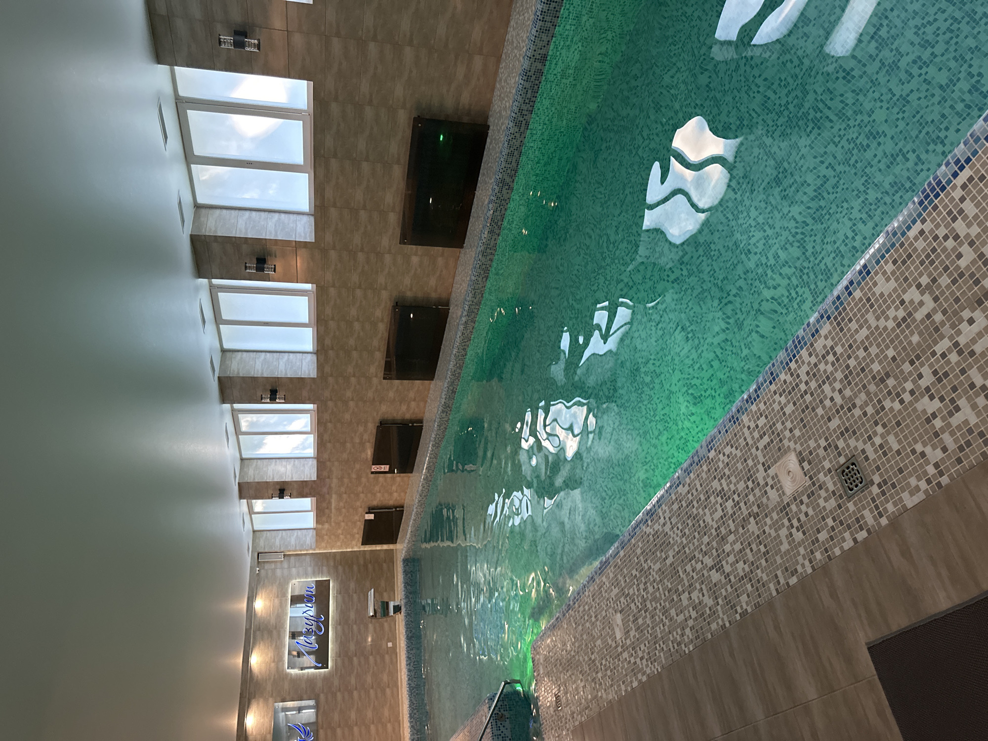 An indoor swimming pool facility is depicted in a long, rectangular pool filled with green-tinted water. The pool is illuminated by underwater lights, creating a vibrant glow. The pool edges are lined with a mosaic of small, square tiles in shades of beige and grey, with a blue border. To the left, a tiled walkway leads to the pool, with a dark brown mat on the floor.
The walls surrounding the pool are tiled with larger, beige rectangular tiles. Along the wall above the pool, a series of windows with frosted glass are evenly spaced. Two black wall sconces are mounted on the wall between each set of windows.
On the wall at the far end of the pool, a neon sign with blue lettering that reads "Лазурный" (Lazurnyy, meaning Azure or Lazuline) is illuminated. To the left of the sign, another mirror with a decorative bird motif is visible. Several dark, rectangular panels are mounted on the wall above the pool, possibly for ventilation or heating.
The ceiling is smooth and white, with recessed lighting panels. The overall impression is of a modern, clean, and well-maintained indoor pool area. The location is identified as Unknown, Unknown.
