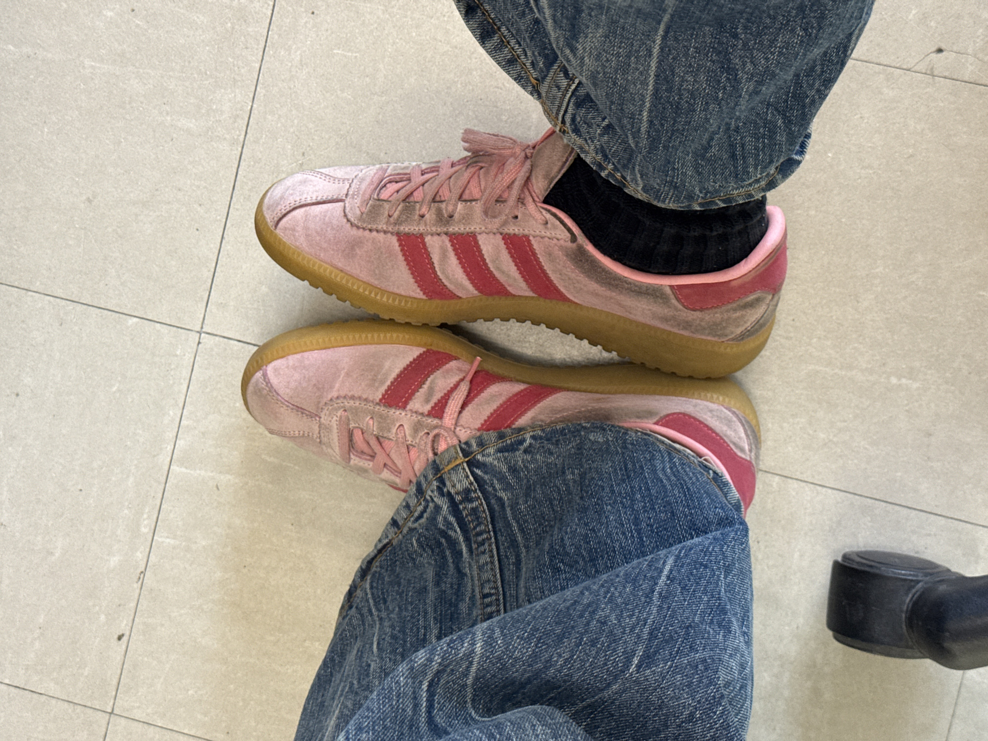 The image shows a person's legs and shoes, likely indoors on a light gray tiled floor. The person is wearing blue jeans and pink Adidas-style sneakers with red stripes and brown soles. Black socks are visible. A black office chair caster is partially visible in the lower right corner. The location is indeterminate as it does not provide visual elements indicating Montreal, Canada.