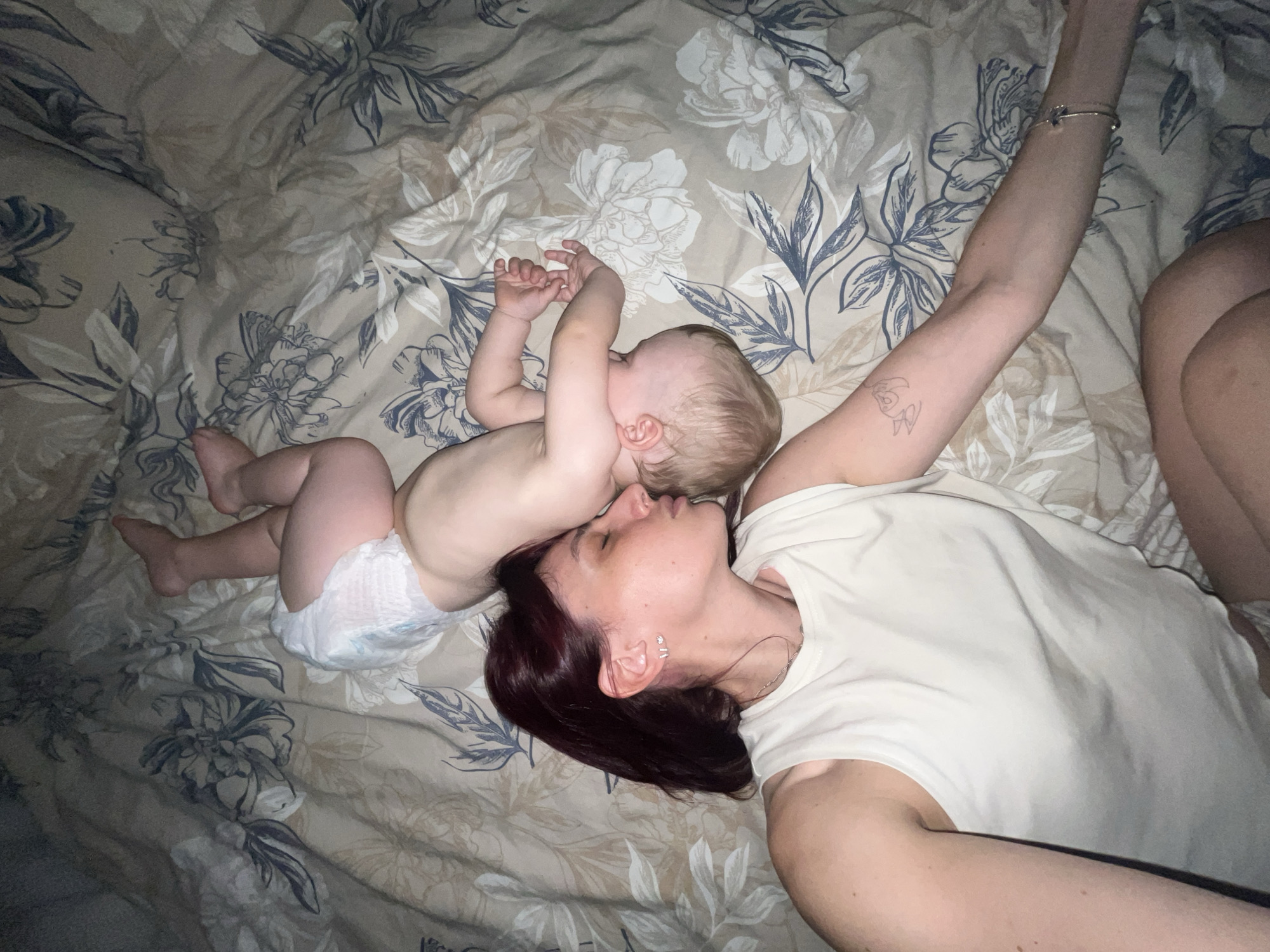 The image shows a mother and her baby lying on a bed with a floral-patterned comforter. The mother, with dark hair, is wearing a white tank top and has a bracelet on her left wrist and a tattoo on her arm. The baby, who is wearing a diaper, is nestled close to the mother, their heads touching. The scene appears to be indoors, in a bedroom, likely during nighttime or in low light conditions. The mood of the image is intimate and peaceful, suggesting a moment of rest or affection between mother and child. There is no visible text in the image.
