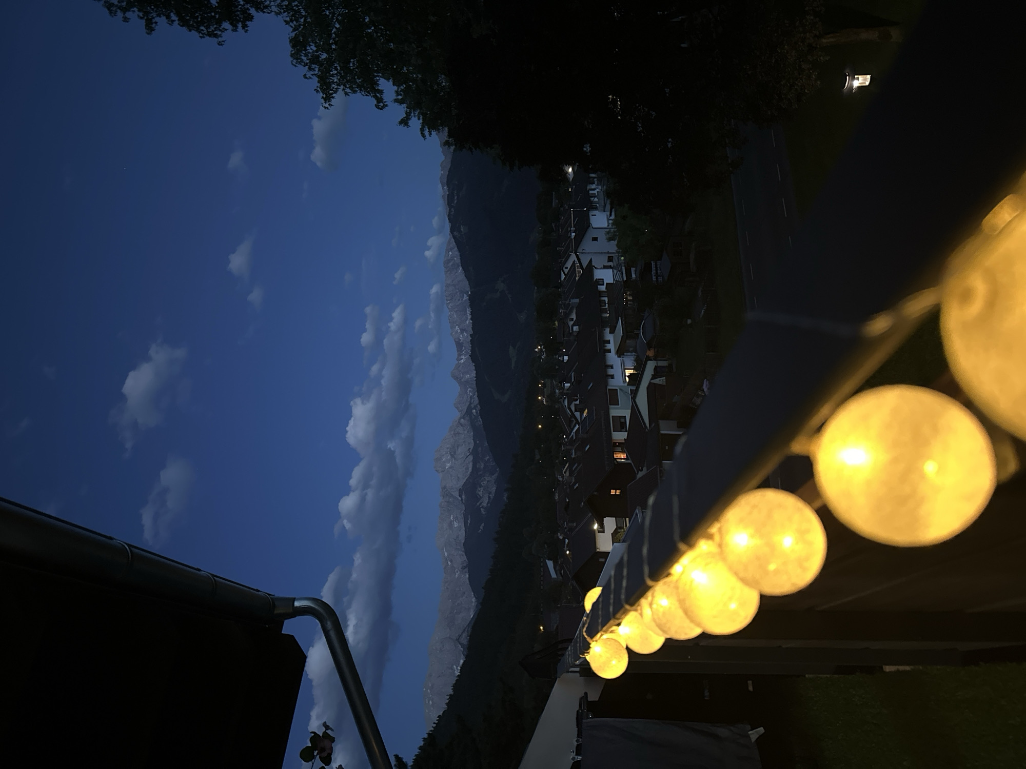 The image depicts a twilight scene in Garmisch-Partenkirchen, Germany, viewed from a balcony or elevated viewpoint. The main subjects are a string of warm, yellow orb lights that extend across the foreground, providing a cozy and inviting ambiance. In the background, a village with white houses and dark roofs is nestled at the base of a majestic, rugged mountain range that dominates the skyline. The sky is a deep blue, transitioning from twilight to night, with scattered white clouds.
The time of day is clearly evening or early night, indicated by the dark sky and the illumination from the string lights and the village windows. The weather appears clear, with the mountains visible in sharp detail against the darkening sky. The presence of the mountains suggests a mountainous or alpine region, consistent with the location context of Garmisch-Partenkirchen.
Notable details include the juxtaposition of the man-made lights and structures with the natural grandeur of the mountains. The angle of the shot, with the string lights in the foreground and the landscape receding into the distance, creates a sense of depth. The image evokes a feeling of peace and tranquility, possibly a moment of relaxation enjoyed on a balcony after a day of activity. There is no visible text in the image.