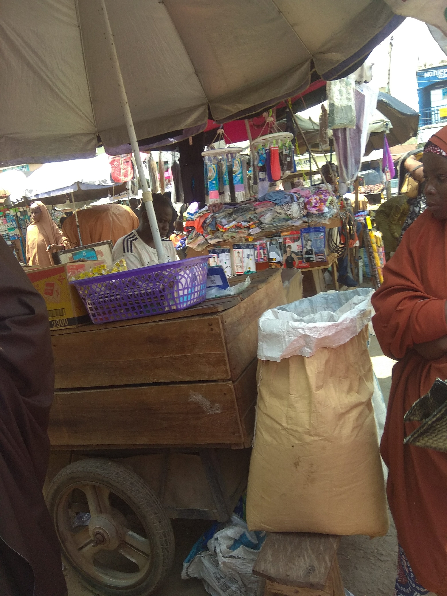 The image depicts an outdoor market scene in Jalingo, Nigeria, likely a street market or a section of a larger bazaar. A large, grey umbrella shades a stall where goods are displayed and sold. In the foreground, a wooden cart with a visible wheel suggests mobility within the market. A substantial sack, light brown in color, sits prominently, possibly filled with goods for sale or transport.
Several individuals are present. To the right, a person wearing an orange headscarf and an orange robe is partially visible, facing away from the camera. In the center, a person wearing a white t-shirt with dark stripes is seated behind a display of items, looking downwards. Further back, another individual in a light-colored hijab stands near a stall laden with goods, including what appears to be fabrics or clothing. Behind them, other market stalls and structures are visible, creating a dense and active commercial environment.
The goods on display include various packaged items, such as what look like underwear or hosiery packs, and a purple basket filled with smaller items, possibly toiletries or accessories. The general atmosphere is that of a busy, informal marketplace. The lighting suggests it is daytime, with strong sunlight casting shadows.