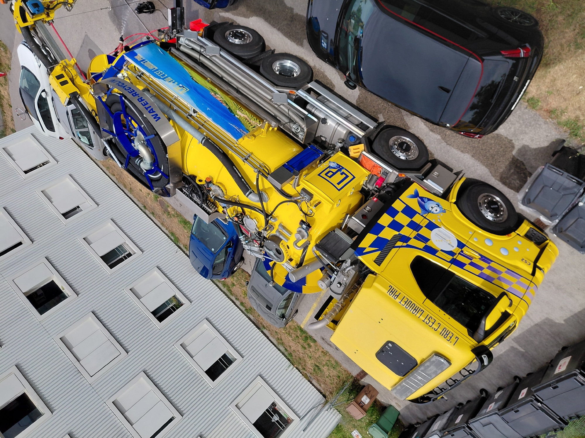 The image shows a yellow and blue vacuum truck parked in front of a corrugated metal building with white-framed windows. The truck has a large tank and various hoses and equipment. A black car is parked to the right of the truck, and a silver car and a blue hatchback are parked behind it. The scene appears to be in an industrial or commercial area, possibly a parking lot. The building's design and the presence of several vehicles suggest an urban or suburban setting. The overall impression is a still-life scene, with no humans or animals visible and no overt actions taking place. The location of Ecublens, Switzerland is not evident from the visual information alone, but the image is consistent with a location in that country.