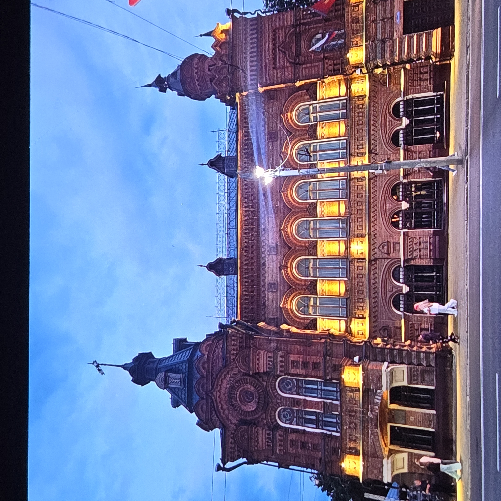 The image depicts the historic building of the Vladimir Museum of Local History, located in the city of Vladimir, Russia. The architecture is characterized by red brick construction with ornate detailing, featuring arched windows illuminated by warm artificial light. The building has multiple towers and spires, one of which is topped with a weather vane. The sky above is a twilight blue with scattered clouds. In front of the museum, a sidewalk is visible with several people walking. One individual in the foreground is wearing a pink headscarf and white clothing. The street in front of the sidewalk shows a white line marking and appears to be an asphalt road. The overall scene captures the building at dusk or early evening.