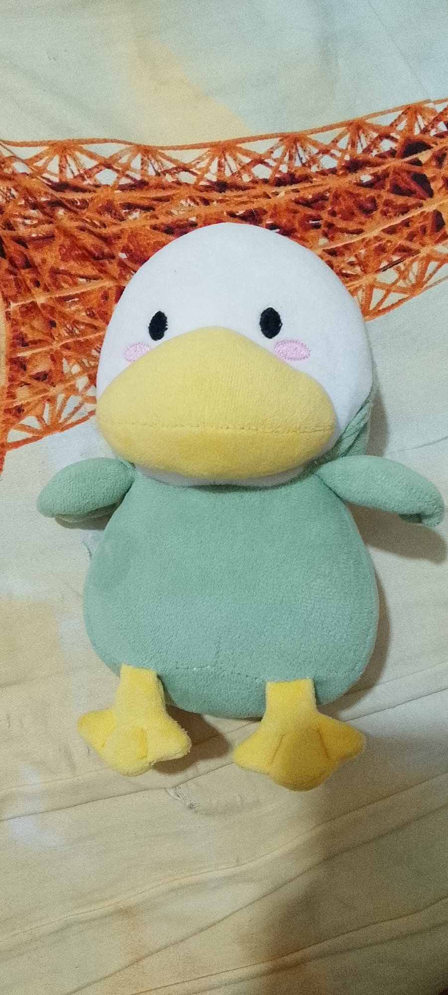 A plush toy duck with a white head, yellow beak, green body, and yellow feet is placed on a light-colored fabric background with a pattern of orange structures. The duck has black eyes and pink blush marks on its cheeks. The background fabric appears to be a blanket or bedspread. The image is taken from a slightly elevated angle, looking down at the toy. There is no indication of the location being in Singkawang, Indonesia.