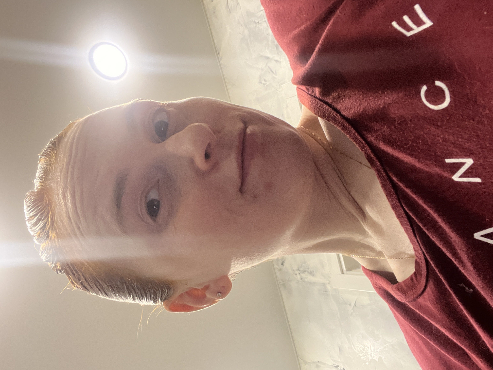 A person is positioned close to the camera, appearing in a low-angle perspective. The person has fair skin, short blond hair, and is wearing a maroon t-shirt with white lettering. A thin gold necklace is visible around the neck. The location appears to be an indoor space with a white or light-colored wall. A light fixture is visible above the person. The photograph does not indicate the location is in the city of Chaikovsky, Russia.