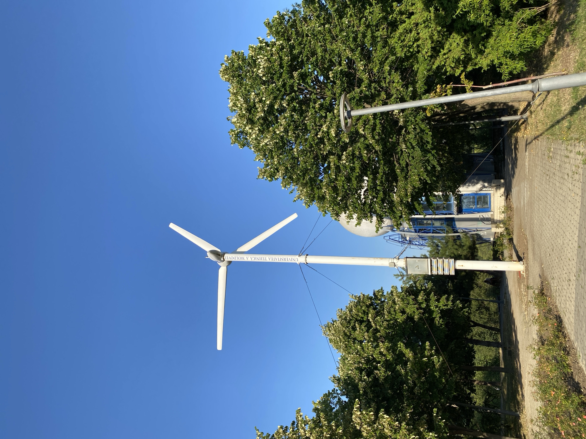 The image depicts an outdoor scene with a small wind turbine prominently positioned in the foreground. The turbine is white and has three blades, with vertical text visible on its pole. Behind the turbine, a large spherical structure, possibly a radar dome or observatory, is visible, along with a building featuring blue-tinted windows and doors. Lush green trees with lighter-colored foliage surround the area, and a paved pathway leads through the scene. A lamppost stands to the right of the pathway. The sky is clear and blue. This location is identified as the Technical University of Moldova in Chișinău, Moldova, specifically near the Faculty of Energetics and Electrical Engineering, where the university's wind turbine is located.