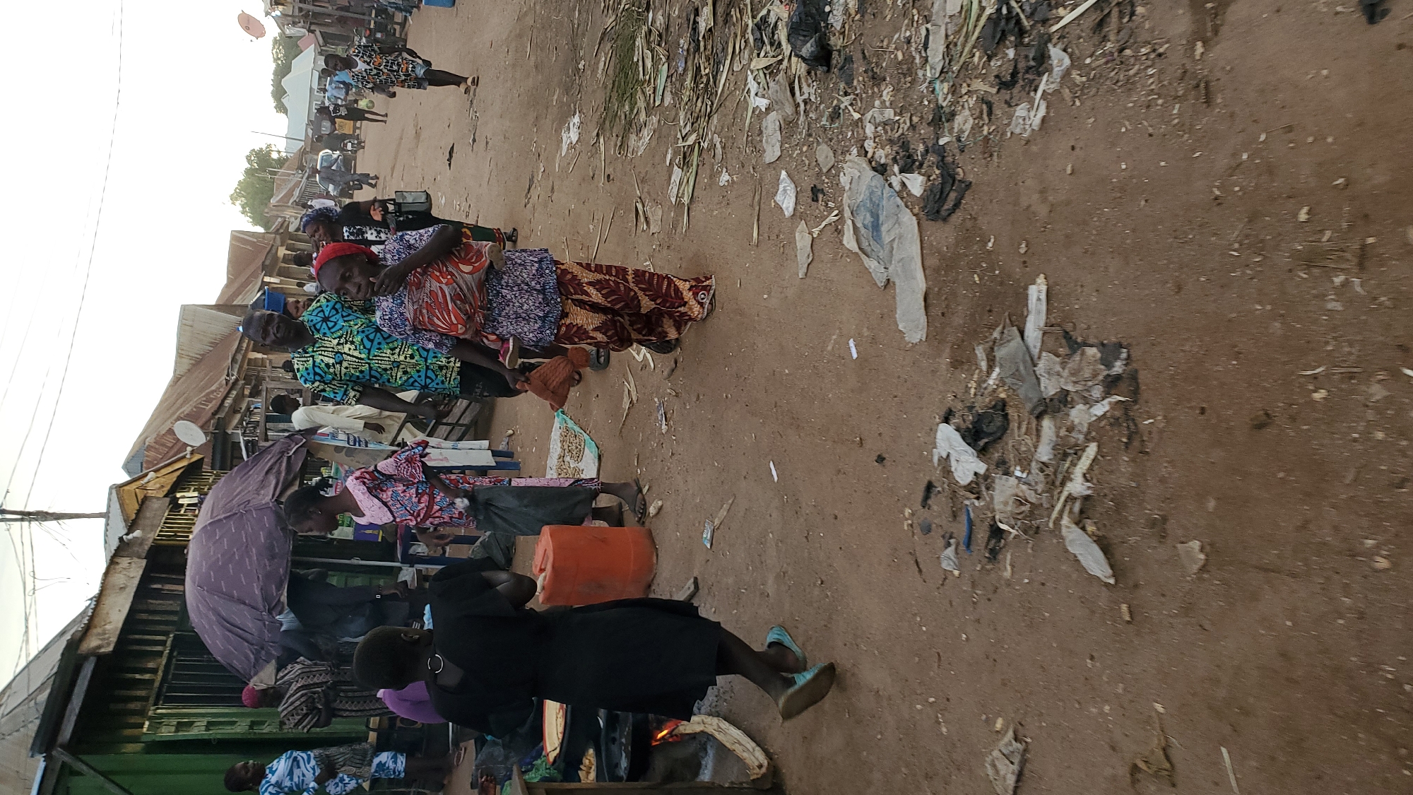 In Jalingo, Nigeria, a dirt street scene unfolds with a collection of open-air market stalls lining one side. A large, dark purple umbrella partially shades a stall where a woman in a patterned dress is visible. Several people are present, including women in colorful traditional clothing, some carrying children. One woman in a black dress with light blue shoes walks away from the camera, toward a cooking area where a fire is visible. An orange plastic drum stands near the market stalls. The street is unpaved and littered with debris, including plastic bags, food scraps, and other refuse. In the background, more market stalls and buildings are visible, along with people and some motorbikes further down the street. The sky is overcast, suggesting it might be late afternoon or early morning.