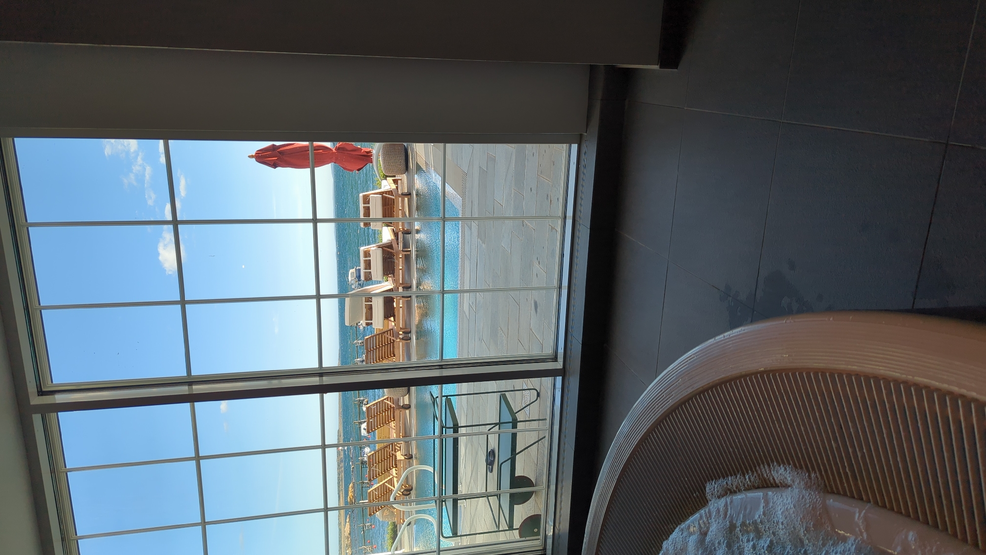 The image depicts a view from indoors, looking through a large window with a grid of panes, out towards a body of water. In the foreground, a portion of a circular, ribbed, tan-colored structure, likely part of a hot tub, is visible on the left. The floor inside appears to be dark tile.
Through the window, a swimming pool with clear blue water is in the mid-ground. Around the pool, several wooden lounge chairs are arranged. To the right of the pool, a red umbrella is closed and upright, with a small, round, textured ottoman nearby. Beyond the pool and lounge chairs, a body of water, possibly the sea, stretches to the horizon under a bright blue sky with scattered white clouds. The coastline in the distance appears rocky and somewhat barren. The scene is likely a resort or hotel with outdoor amenities.
The location is identified as Larkollen, Norway. This suggests the view is of the Oslofjord or the Skagerrak. The outdoor setting with a pool and lounge chairs implies a recreational or resort area within Larkollen. The presence of the hot tub in the foreground indicates an indoor spa or relaxation area connected to the outdoor view.