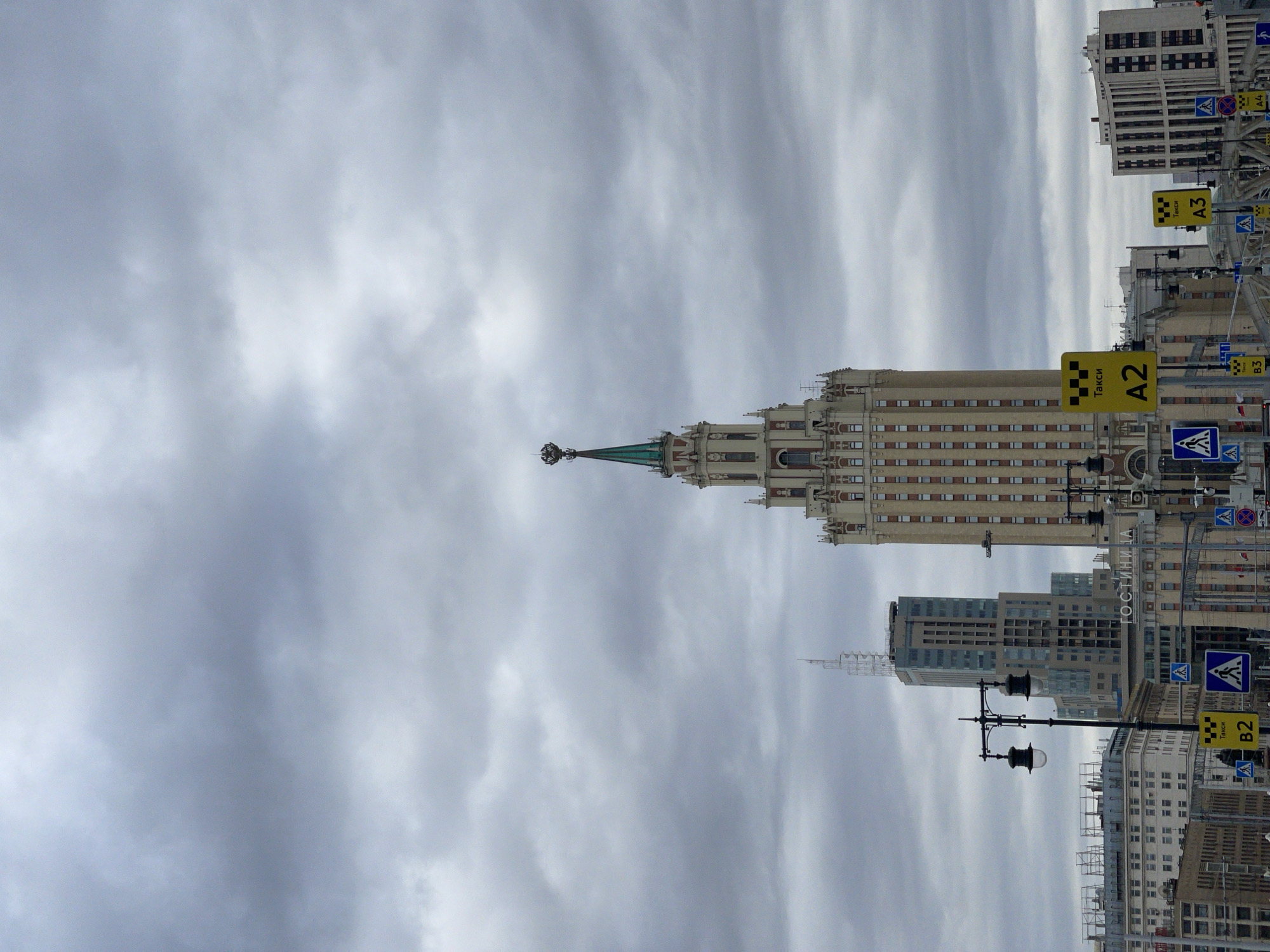 The image shows a cityscape in Moscow, Russia under an overcast sky. The Kudrinskaya Square Building, one of the "Seven Sisters" Stalinist skyscrapers, is prominently featured. Various buildings and street signs, including yellow taxi signs labeled A2, A3, and B2, are visible. The lighting suggests daytime, but the overcast sky results in a muted color palette. A street lamp is positioned in the lower right, along with pedestrian crossing signs. The environment appears urban with no people visible.