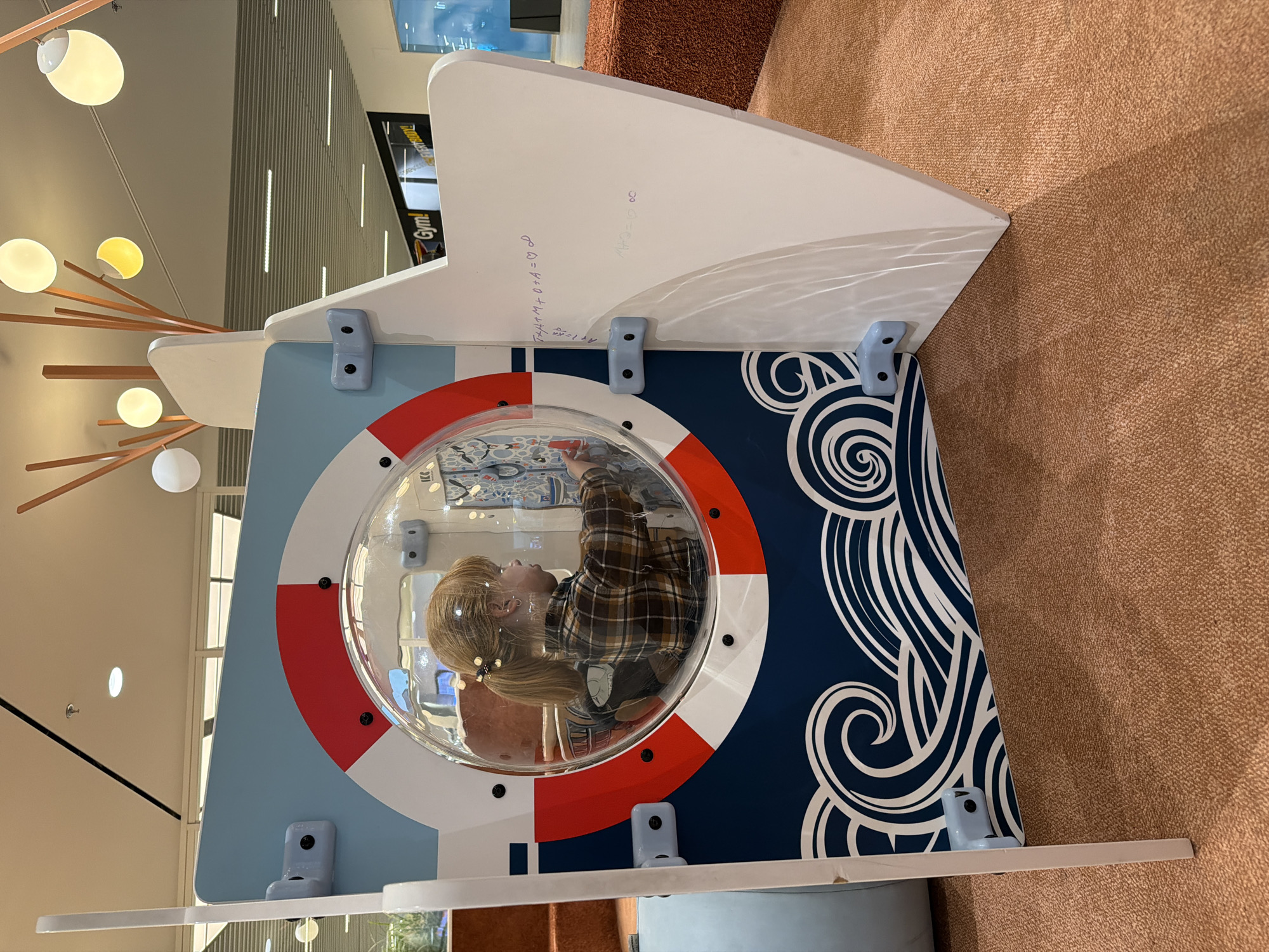 A young child with blonde hair in a ponytail, wearing a brown and black plaid shirt, is looking at a colorful interactive panel through a round, clear plastic window on a white and blue boat-themed structure. The structure has a red and white life preserver graphic around the window and a wave pattern on the lower portion. The child's hands are visible, interacting with something inside the structure. The background shows an indoor environment with a patterned carpet, seating, and overhead lighting. The structure is located in Tallinn, Estonia, within a recreational area, possibly a shopping center or play space.