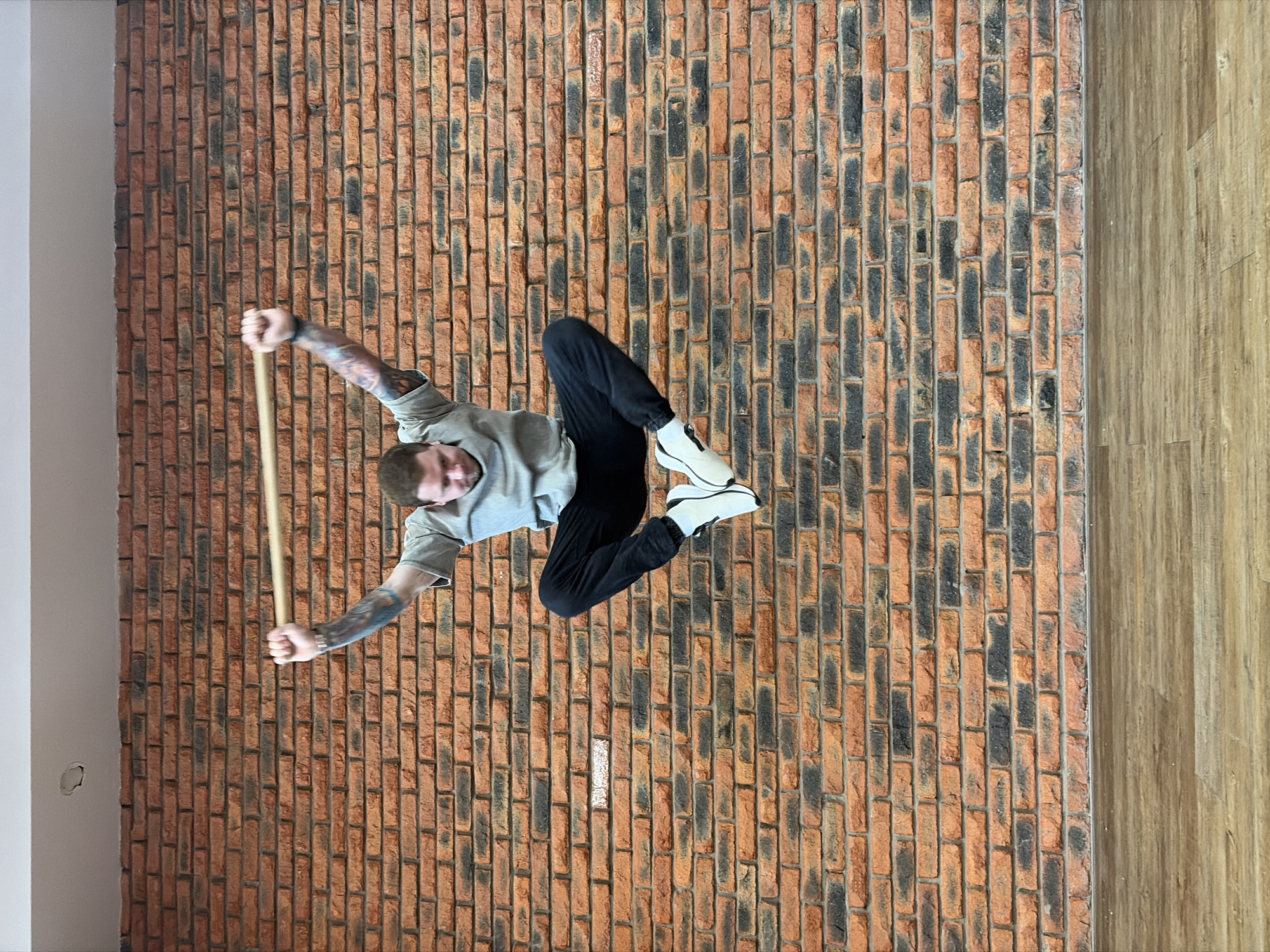The image, rotated 90 degrees counter-clockwise, presents a man seemingly suspended horizontally against a rustic brick wall, holding a wooden stick. The man, appearing to be in his 20s or 30s, has short brown hair, a beard, and visible tattoos covering his arms. He wears a grey t-shirt, black joggers, and white sneakers with black soles and zipper details. He looks towards the camera with a neutral expression, his body angled with bent legs, suggesting an acrobatic pose or physical feat.
The primary background is a textured brick wall with varying shades of red, orange, and dark grey bricks, appearing as vertical columns due to the image rotation. To the right, a wooden surface, resembling a floor or paneling with visible grain and plank lines, runs parallel to the brick wall. To the left, a plain white wall or pillar is partially visible.
In reality, the scene likely depicts the man lying on his back on a wooden floor, positioned next to a brick wall. He holds the wooden stick horizontally above him, and his legs are bent and slightly raised. The image has been intentionally rotated to create an optical illusion of him defying gravity. The setting is an indoor space, brightly lit, presumably during the daytime. No discernible text is visible within the frame. This scene takes place in Chaikovsky, Russia.