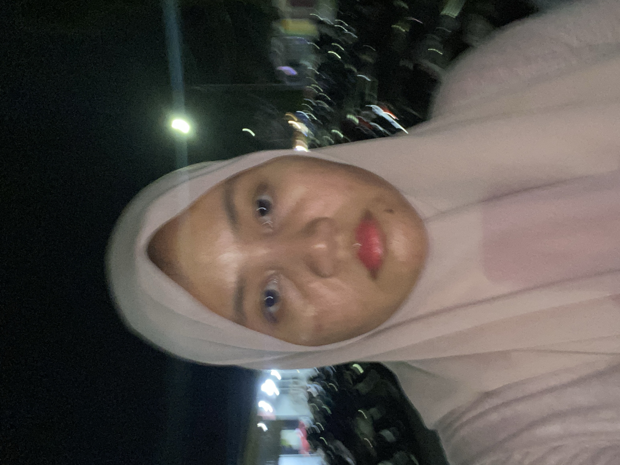 A selfie, taken at night, shows a person wearing a light pink hijab and shirt. Their face is partially illuminated by a street light in the background, revealing skin imperfections. The person has dark eyes and pink lipstick. Behind them, rows of parked motorcycles are visible, with blurred lights indicating a city setting in Banjarbaru, Indonesia.