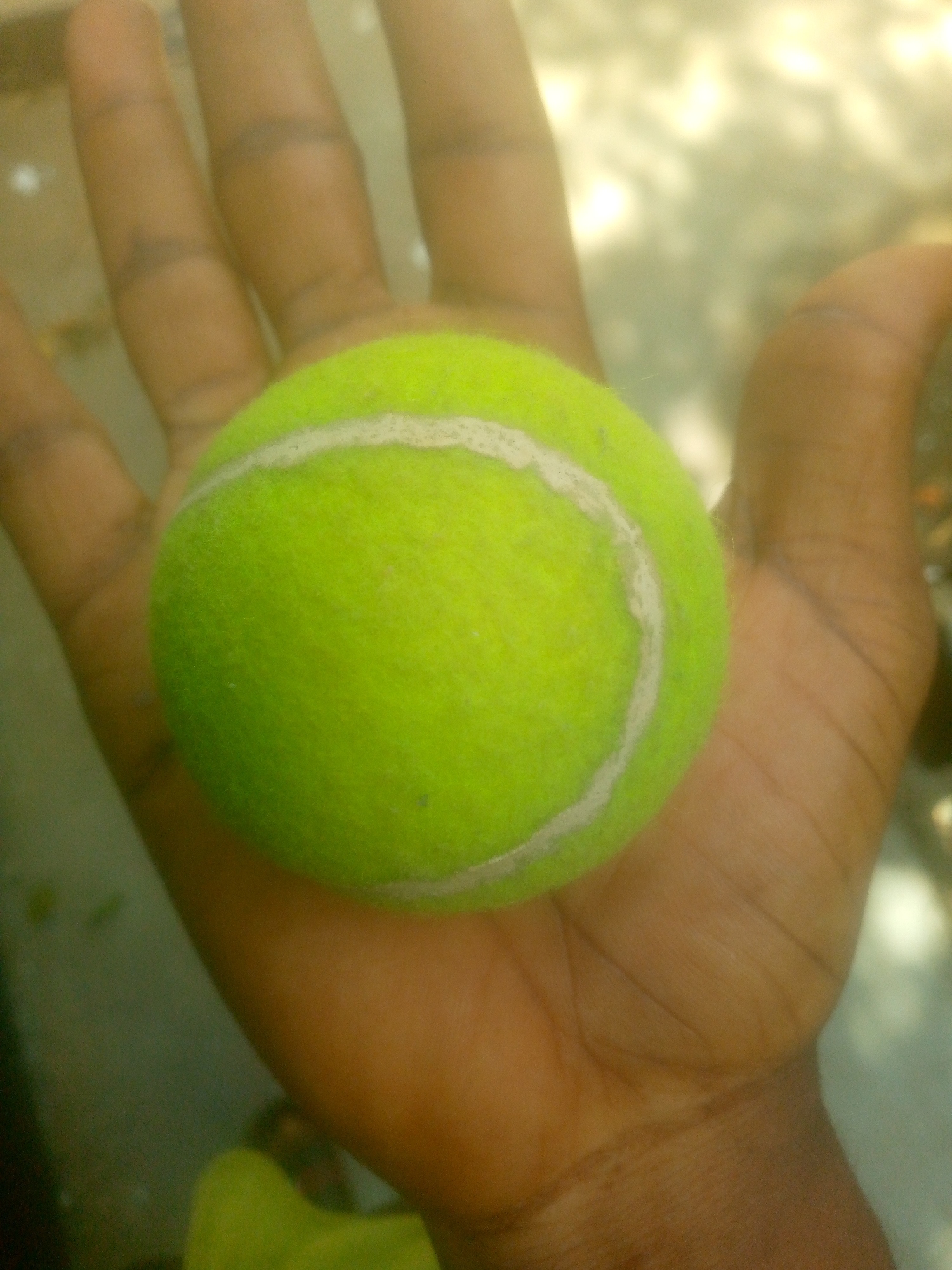 A close-up image shows a human hand holding a bright, fluorescent yellow-green tennis ball. The hand is open, with fingers slightly spread and visible in the upper and lower portions of the frame, providing scale for the ball. The skin tone of the hand is medium to dark brown, and creases are visible on the palm and fingers. The tennis ball is prominently featured in the center, showcasing its characteristic felt-like surface and the distinct white curvilinear seams. The lighting appears bright, possibly from natural daylight, highlighting the vivid color of the ball. The background is completely out of focus, rendered as soft, indistinct blurs of lighter and darker tones, preventing any specific identification of the environment or setting. No discernible activities other than holding the ball are taking place, and no text is visible in the image.