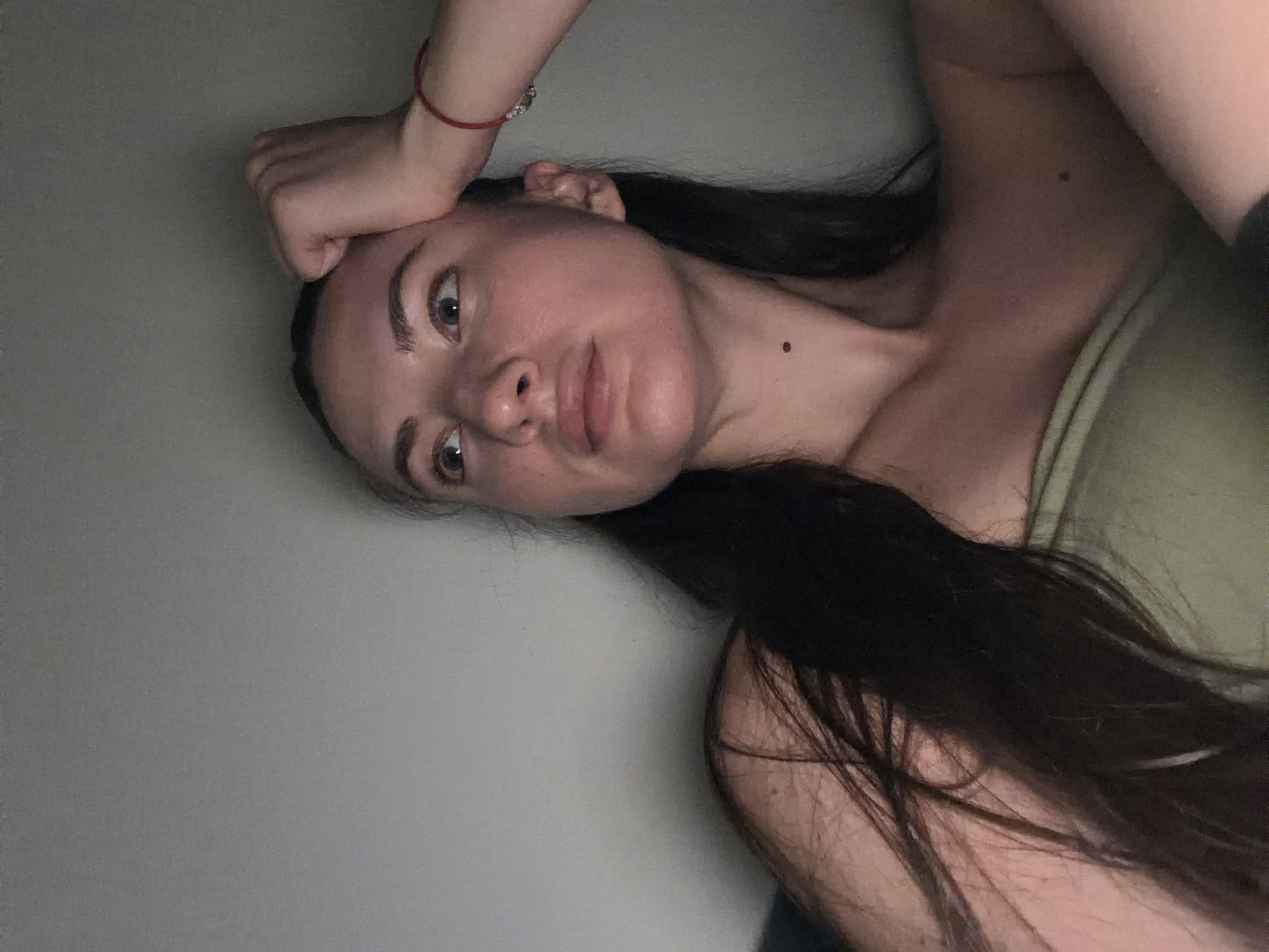 A young woman with long, dark hair is captured in a medium shot, looking upwards and to her left with a pensive expression. Her right hand is resting on her forehead, and a thin red bracelet is visible on her wrist. She is wearing a light green strapless top. The background is a plain, light gray wall. The lighting is dim, with a noticeable glow on her face and shoulder. There are no discernible elements in the image that suggest a specific location in Sharm El Sheikh, Egypt.
