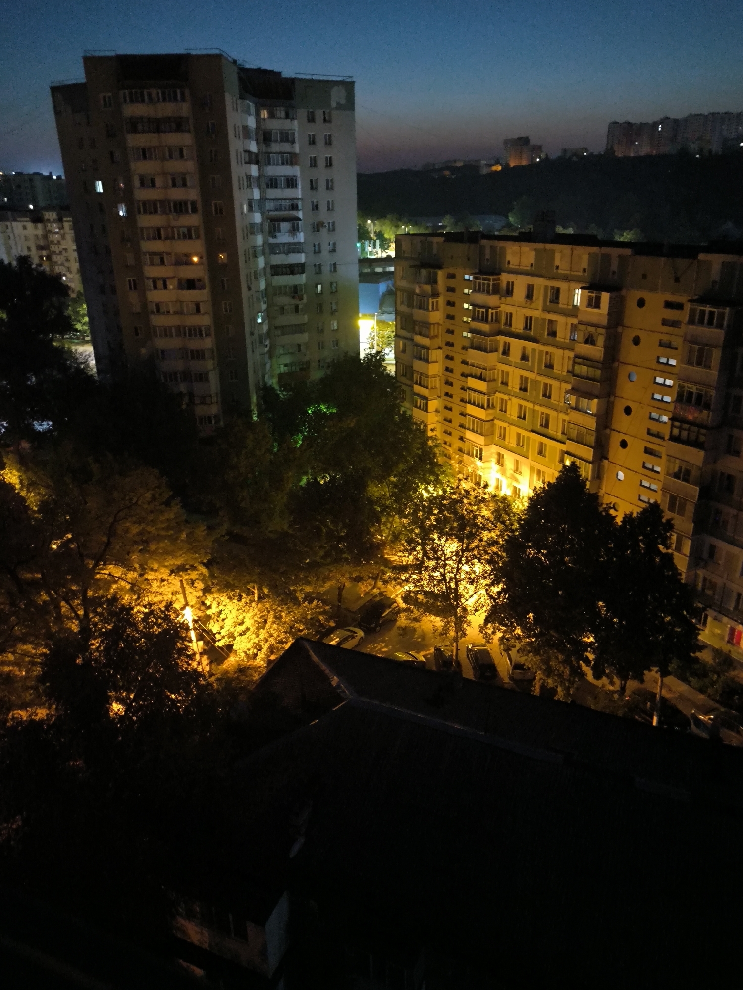 The image depicts an urban nighttime scene from an elevated perspective, likely overlooking a residential area in Chișinău, Moldova. Two prominent apartment buildings are visible, one to the left and another to the right, both illuminated from within, indicating occupancy. The building on the left is taller and appears older, with a grey facade and numerous balconies. The building on the right is also multi-story, with a yellowish-beige exterior, featuring a more regular pattern of windows and balconies.
In the foreground, the dark silhouette of a rooftop obscures part of the view. Between the buildings and the foreground, a cluster of trees is illuminated by artificial lights, casting a warm, orangish glow on their foliage. Streetlights and lights from ground-level structures are also visible, contributing to the overall ambiance of the urban night. The sky above is a deep, dusky blue, transitioning to a lighter hue near the horizon where faint lights suggest distant city structures on a gently rolling terrain. Several cars are parked in the illuminated areas, suggesting a typical residential street scene. No individuals are discernible in the image. The overall impression is one of a quiet, possibly late-evening or early-morning, urban environment.