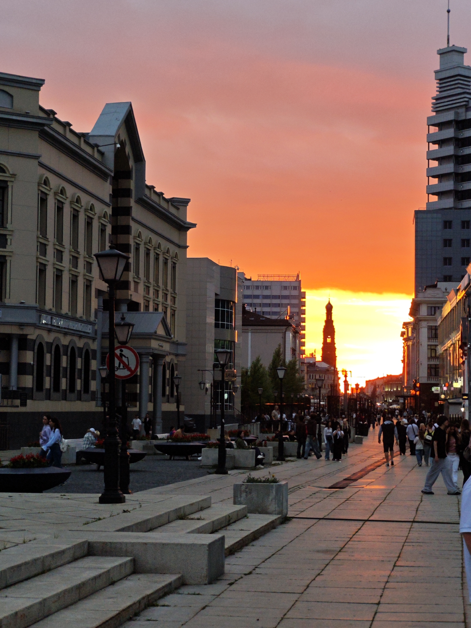 The image depicts a bustling street scene in Kazan, Russia, during a vibrant sunset. The sky is ablaze with hues of orange and pink, casting a warm glow over the city. People are strolling along the wide pedestrian walkway, some in small groups, others alone. The architecture lining the street features a mix of historical and modern buildings, with a prominent tower visible in the distance, silhouetted against the colorful sky. Streetlights are illuminated, suggesting the approach of evening. The atmosphere appears lively and active, with a sense of community as people enjoy the pleasant weather and scenic view. The cobblestone pavement and various architectural elements provide strong location cues for Kazan.