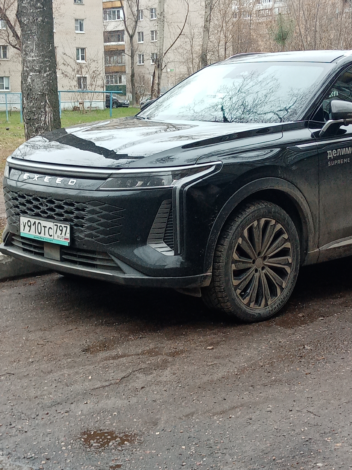 A black Exceed SUV is parked on a wet asphalt surface. The vehicle's front end is visible, showcasing the grille with the "EXEED" emblem, a Russian license plate "Y910TC797," and a distinctive headlight design. The front passenger side wheel is also visible. A building with multiple windows and a bare tree are in the background. The word "Supreme" and a Russian word are visible on the side of the vehicle. This image shows an urban environment; the location is deduced to be Elektrostal, Russia, based on the license plate.