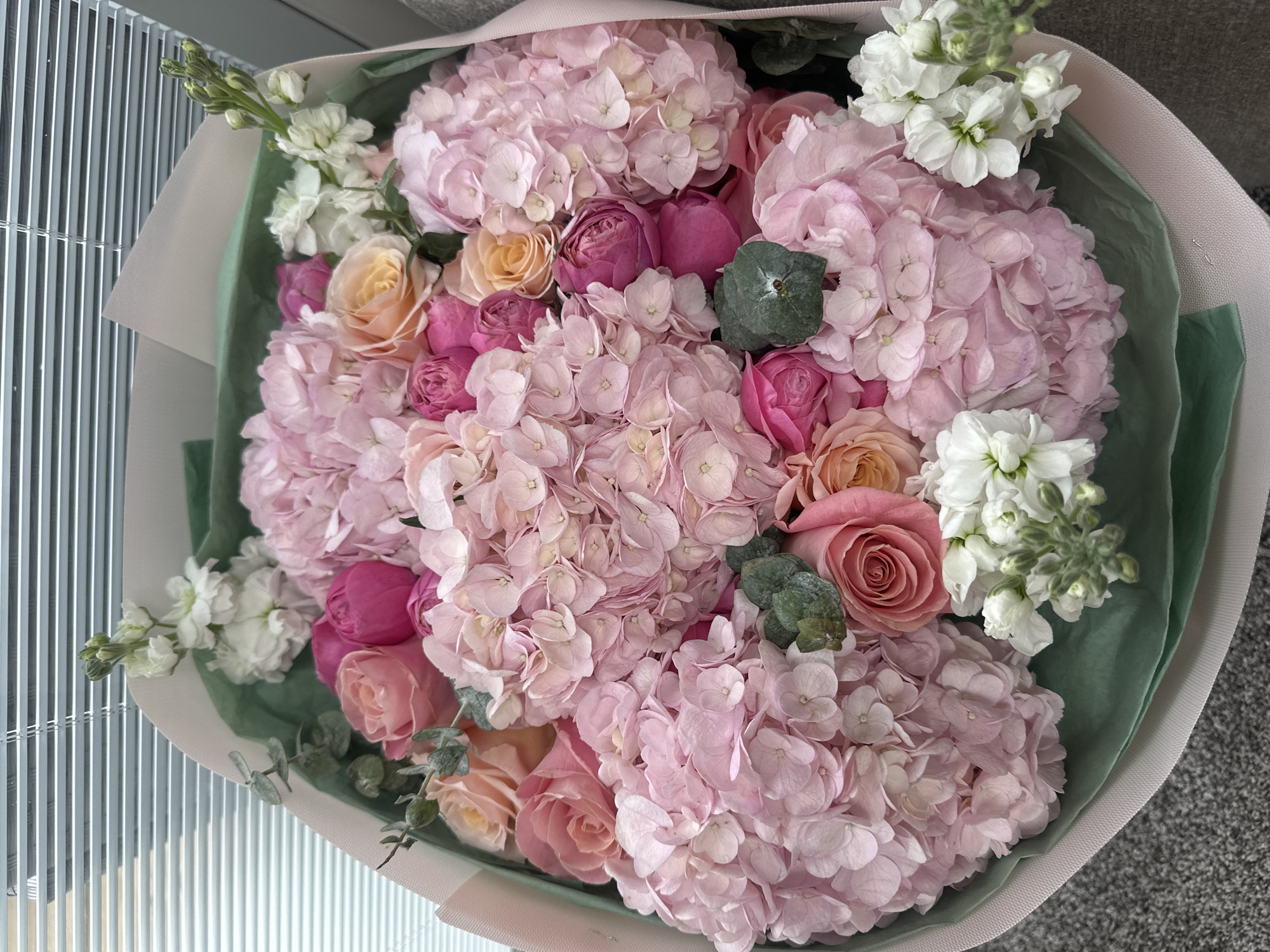 A close-up, overhead shot shows a large bouquet of pink and peach roses and hydrangeas. The flowers are artfully arranged in a light pink and green paper wrap, with some sprigs of greenery interspersed throughout. The bouquet sits on a textured gray surface, possibly carpet, and a light-colored windowsill with horizontal blinds is visible in the upper portion of the image. The lighting suggests an indoor setting.