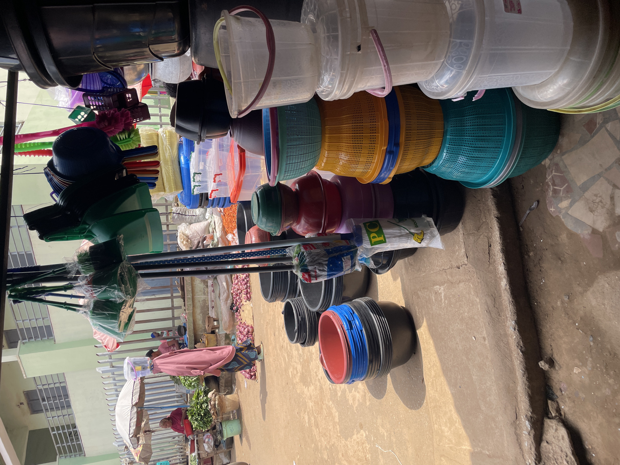 In Akwanga, Nigeria, a bustling market scene unfolds. A woman, draped in a vibrant pink garment and a headscarf, navigates the dusty ground, carrying a clear plastic container filled with water on her head. Beside her, a vendor displays a variety of household items, including brooms, dustpans, and an array of plastic containers of varying sizes and colors, neatly stacked. The market is lined with stalls selling produce, with onions and leafy green vegetables visible. The background features a light green building with a metal railing, hinting at the urban environment. The sunlight casts long shadows, indicating it's likely midday or afternoon.