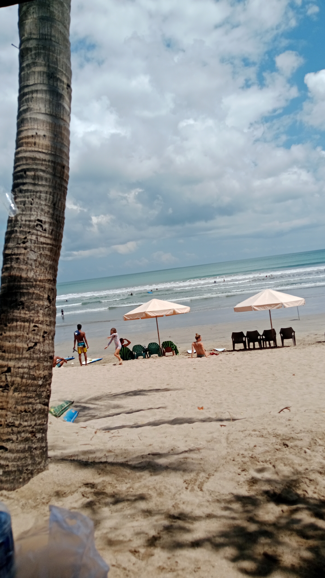 In Kuta, Indonesia, a beach scene unfolds under a cloudy sky with patches of blue. The foreground is dominated by the textured trunk of a palm tree on the left. The sandy beach stretches towards a turquoise ocean with moderate waves. Several people are present: a man in yellow shorts stands on a surfboard near the water's edge, a young girl in white is running on the sand, and a woman in a bikini sits on the sand facing away from the camera. Two peach-colored beach umbrellas provide shade for rows of empty green and brown beach chairs. In the distance, several surfers are visible in the water. The scene captures a typical day at the popular Kuta beach.