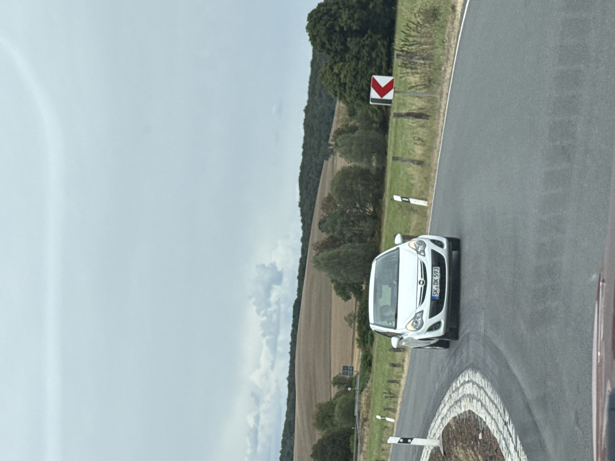 This is a frontal, eye-level, medium shot of a white Opel Corsa driving on a winding rural road in Hohenstein, Germany. The car is centered in the frame and is facing forward. The license plate is visible and reads "SK-DK 593". The road is asphalt with white lane markings. To the left of the road, there is a cobblestone circular island, and on the right side of the road, a traffic sign with a red and white arrow indicates a sharp curve to the left. Beyond the road and fields, a rolling landscape of harvested fields and dense green forest can be seen under a cloudy sky. There are two people visible inside the car, a driver and a passenger. The overall impression is a scenic drive through the German countryside on a cloudy day.