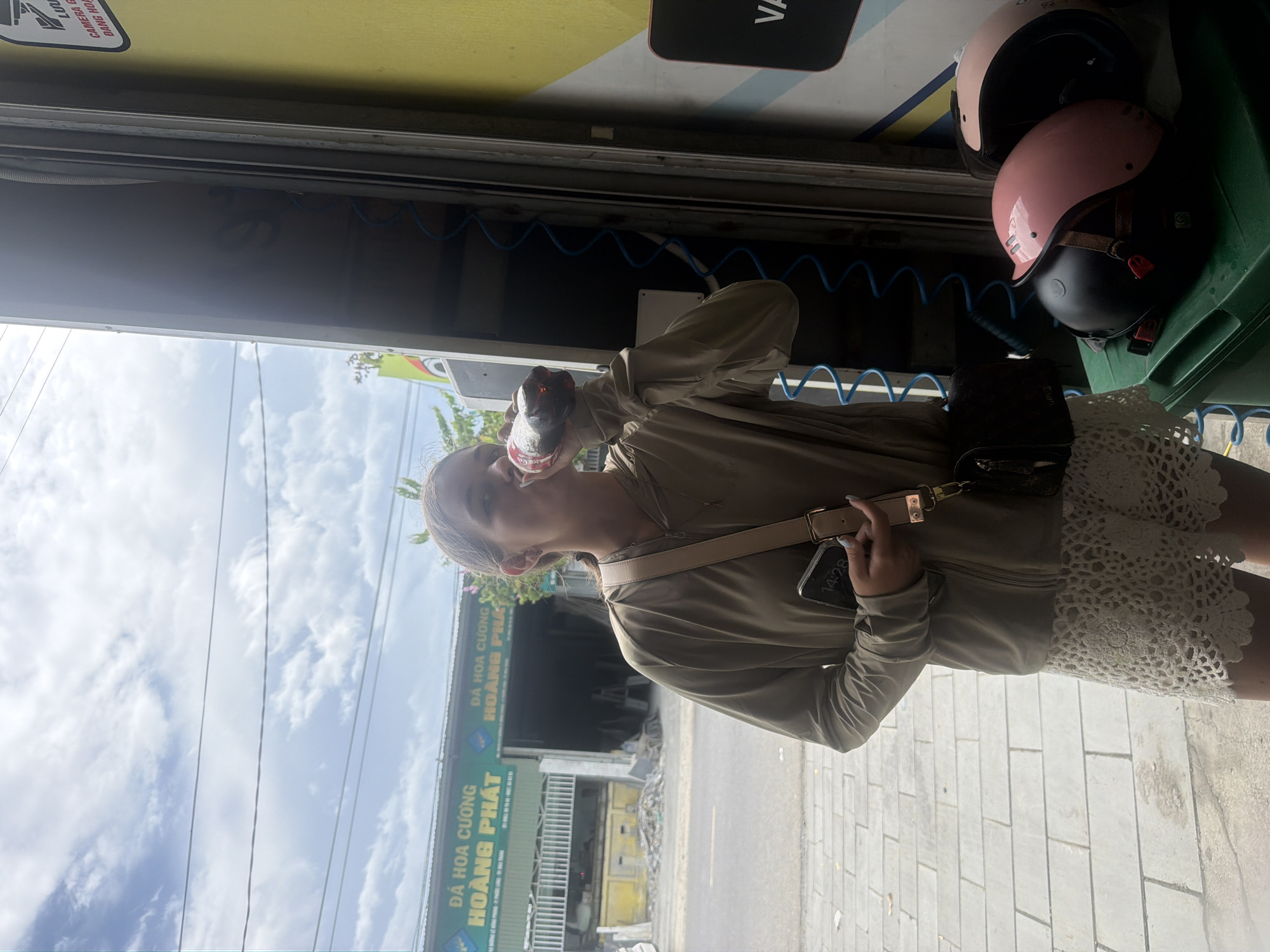 A young woman with blonde hair pulled back drinks from a bottle of soda while standing on a tiled sidewalk. She is wearing a light brown hooded jacket and white crochet shorts. A beige crossbody bag is slung over her shoulder. A smartphone is held in her right hand, displaying the time 14:28. To her right, inside an open service bay or storefront, are two helmets, one black and one pink, placed on top of a green bin. Behind her, a building with green siding and signage in Vietnamese reads "DA HOA CUONG HOANG PHAT." The building appears to be a stone or marble supplier, with large slabs visible inside. The sky above is cloudy and blue, with power lines crisscrossing overhead. The overall scene suggests an outdoor commercial area in Nha Trang, Vietnam.